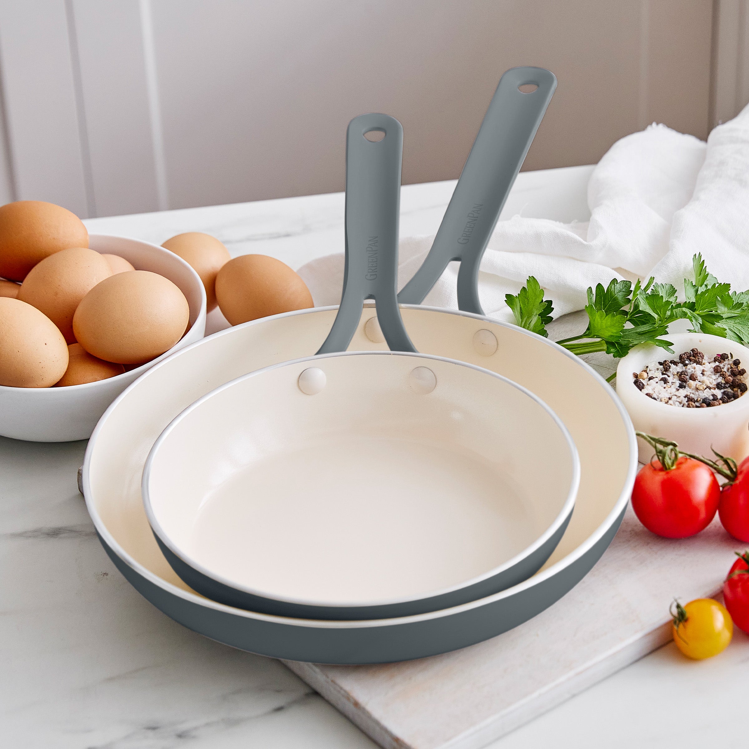 Set of frypans with gray handles on a kitchen counter with eggs and vegetables.