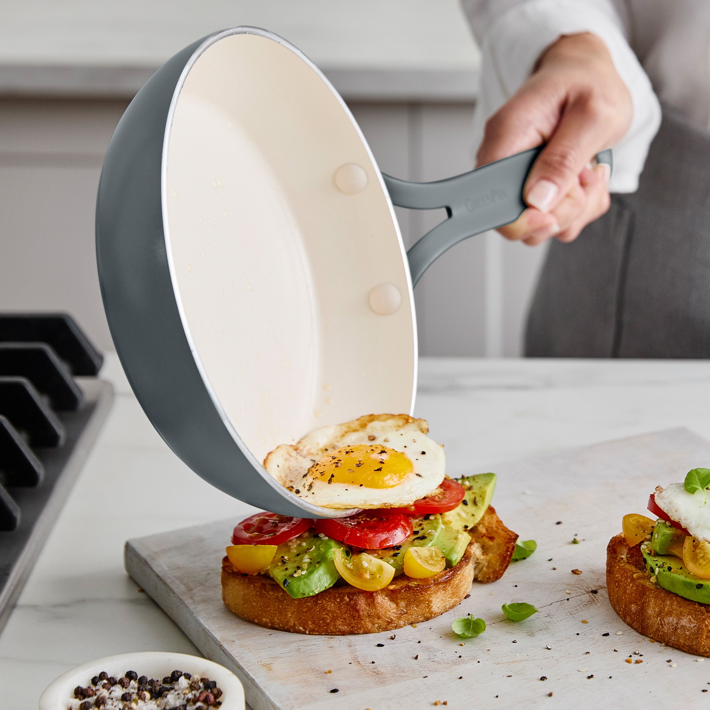 Person using a frying pan to place an egg onto avocado toast on a marble countertop.