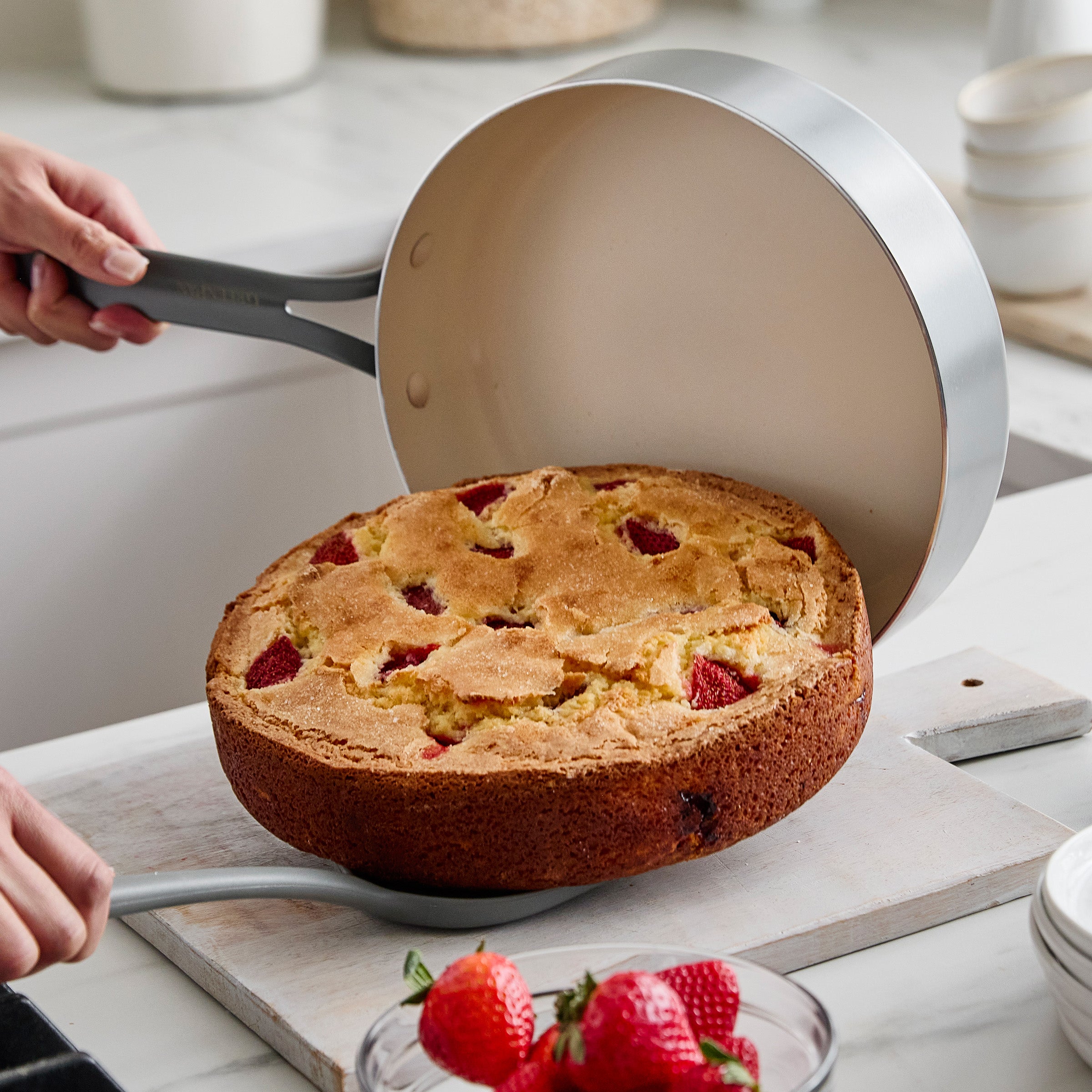 Person lifting a cake out of a round pan on a kitchen counter with strawberries nearby.