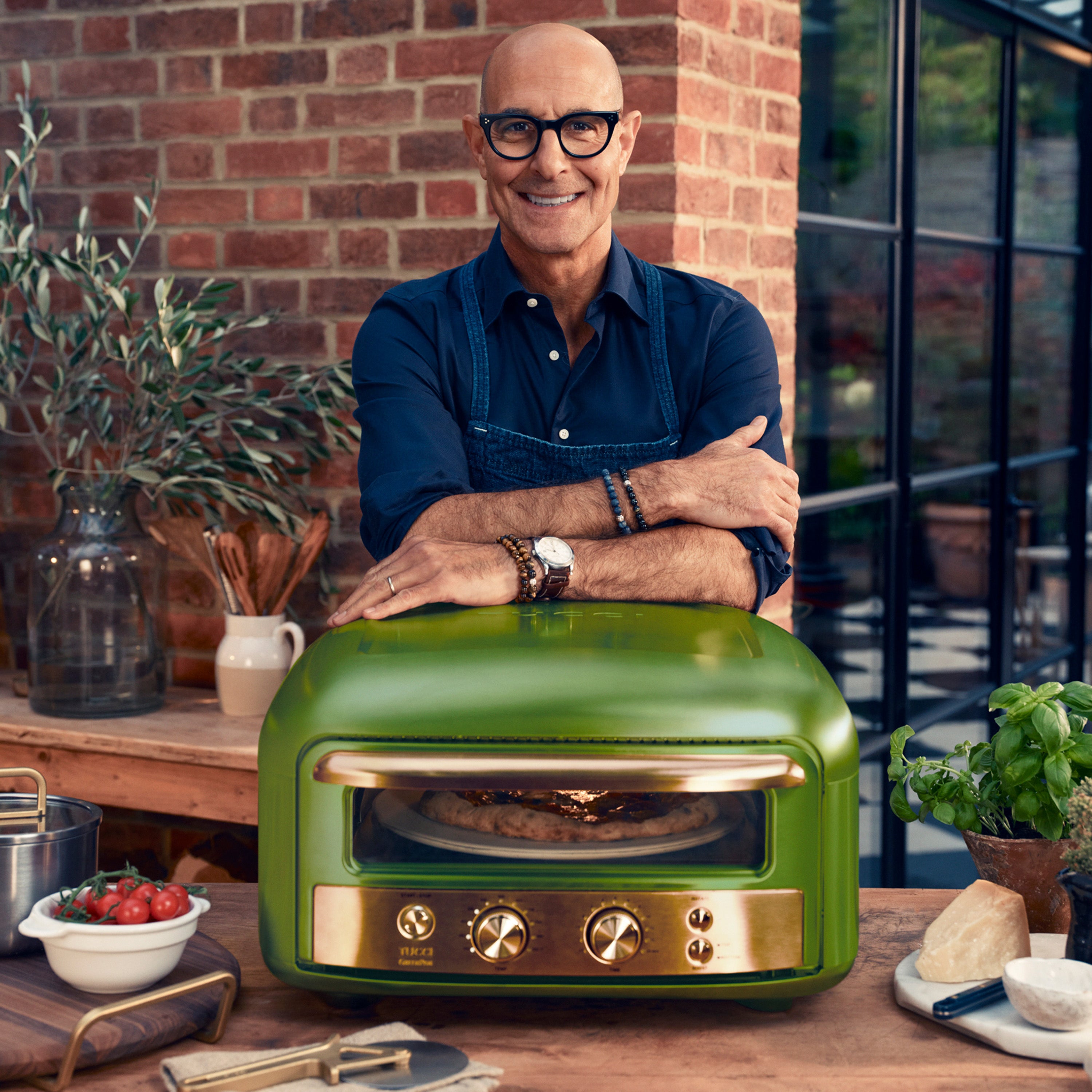 Stanley Tucci standing next to a green pizza oven on a wooden table with plants and kitchen items in the background.