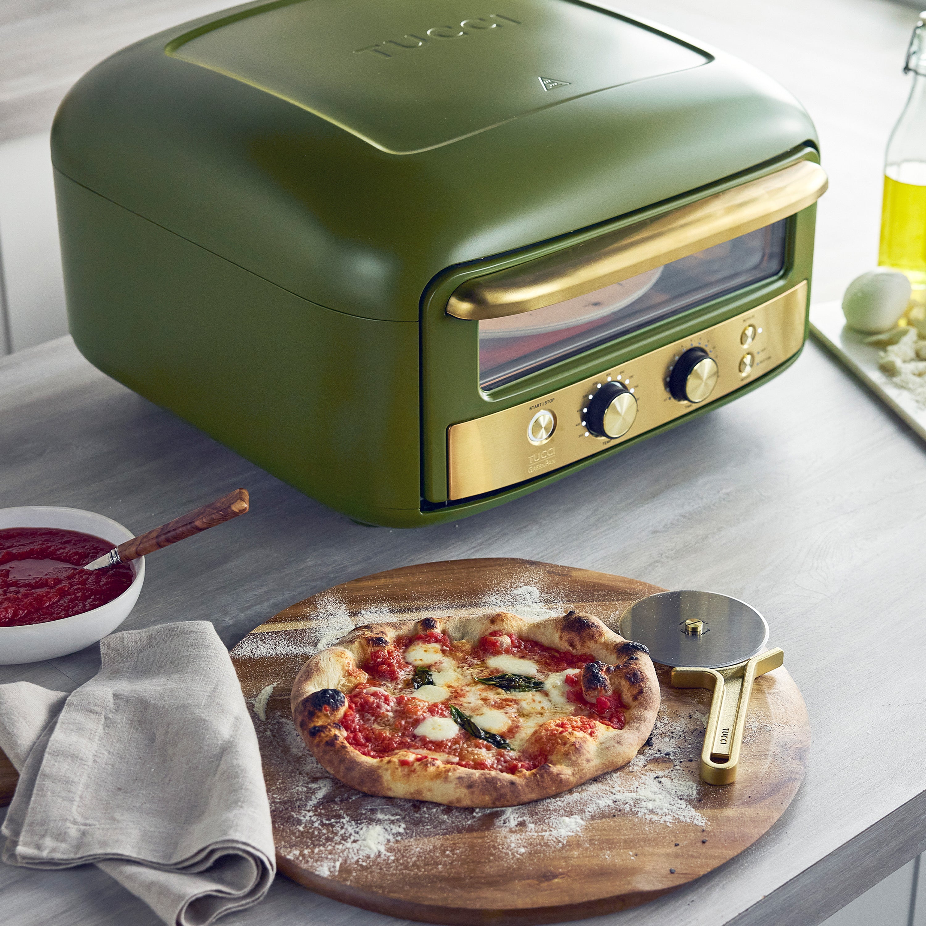 Green pizza oven with a pizza on a wooden board, surrounded by kitchen items.