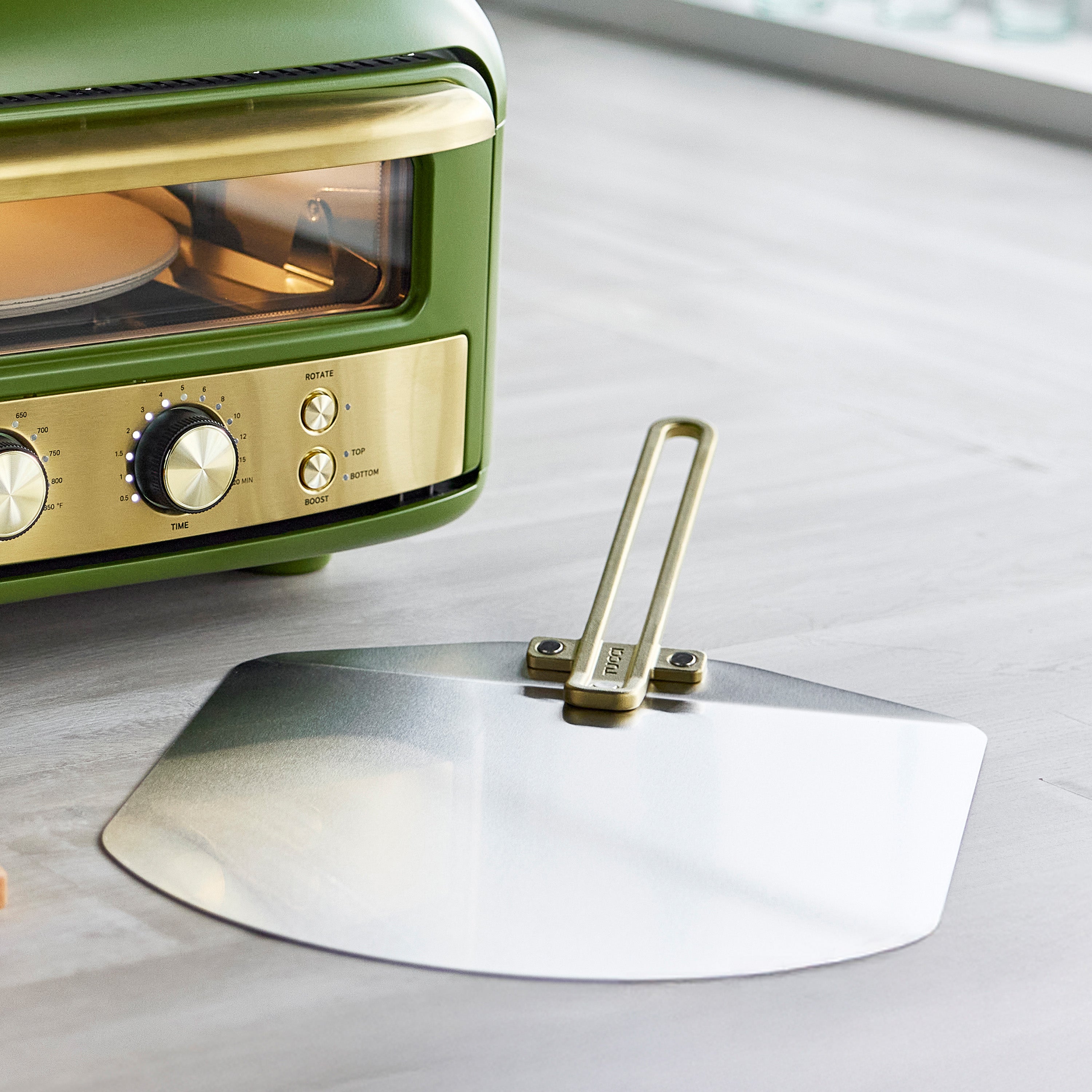 Green and gold pizza oven with a metal pizza peel on a light gray surface