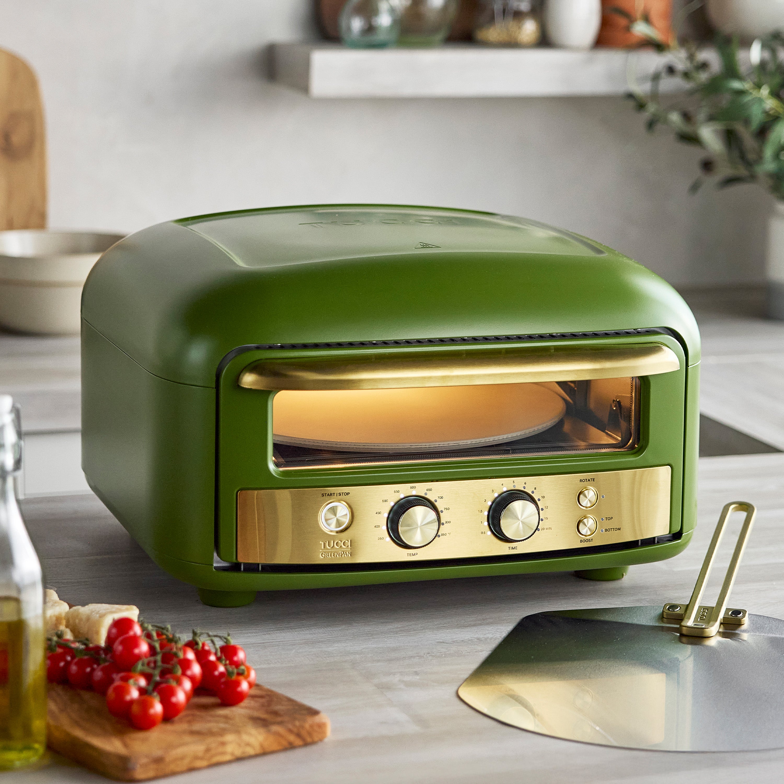 Green and gold pizza oven on a kitchen counter with tomatoes and bread.