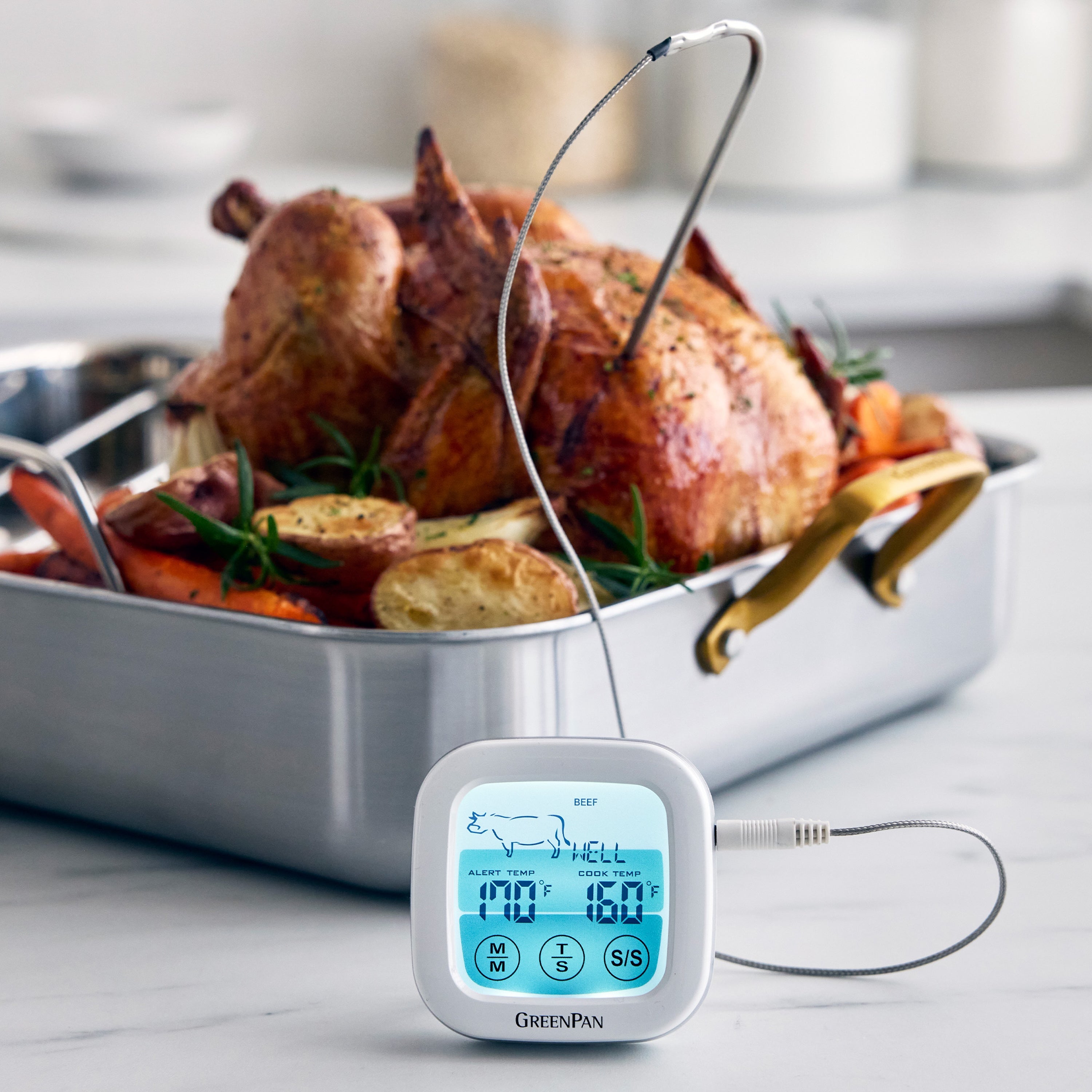 Digital oven thermometer displaying the temperature in front of a roasting pan with chicken and mixed vegetables.