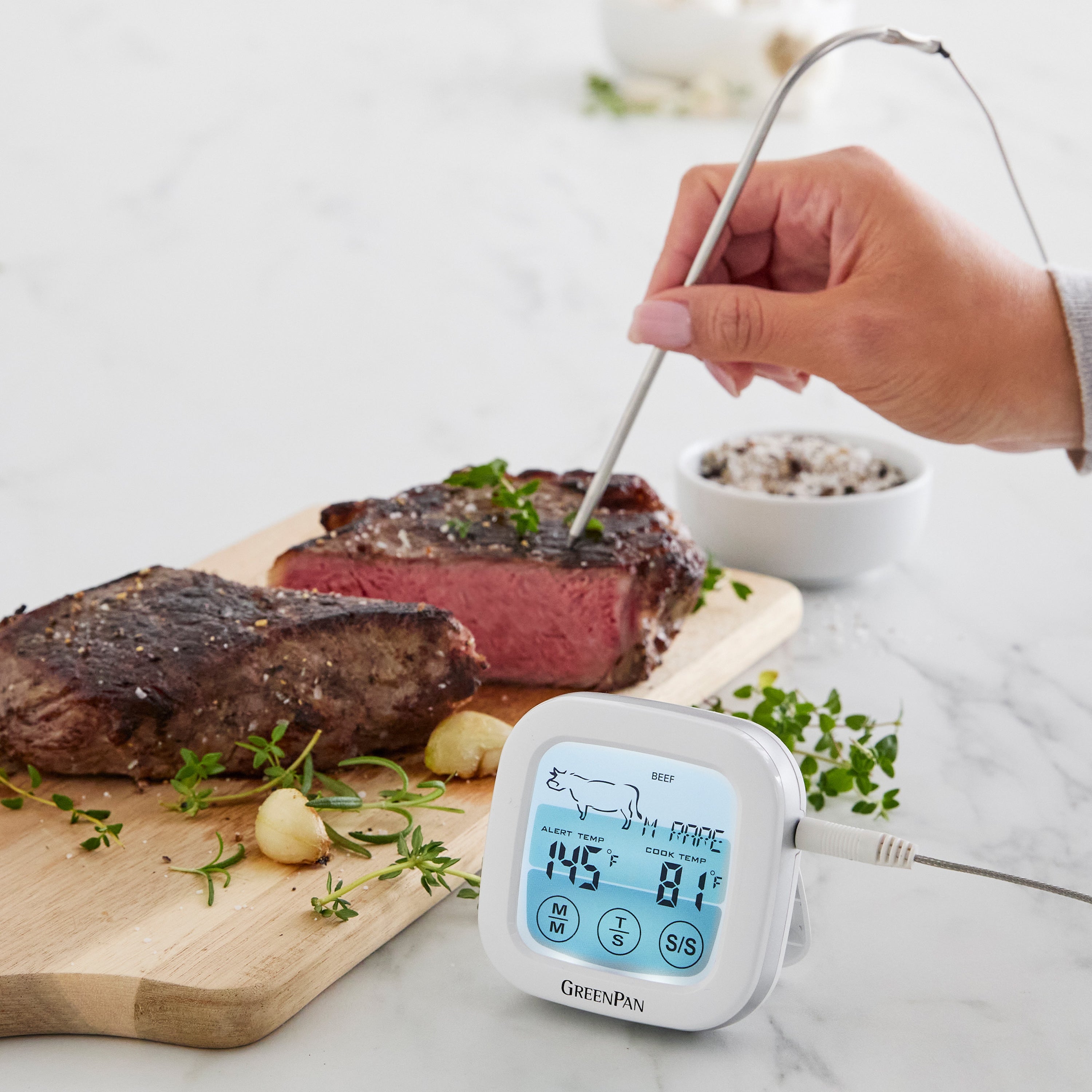 Hand checking steak doneness with AccuRead digital oven thermometer on a wooden cutting board garnished with herbs.