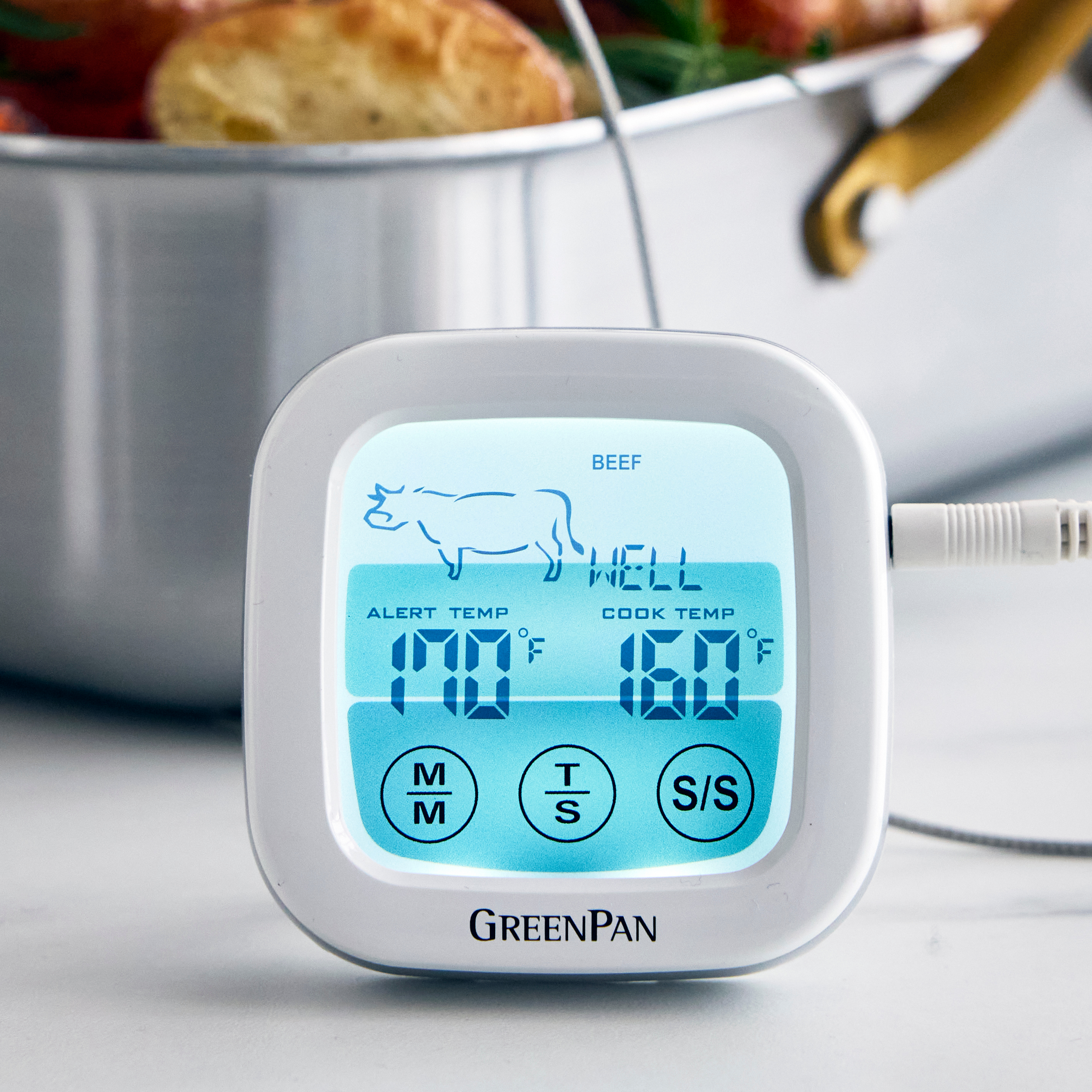 Digital oven thermometer displays beef cooking temps; background shows blurred food in a pan.