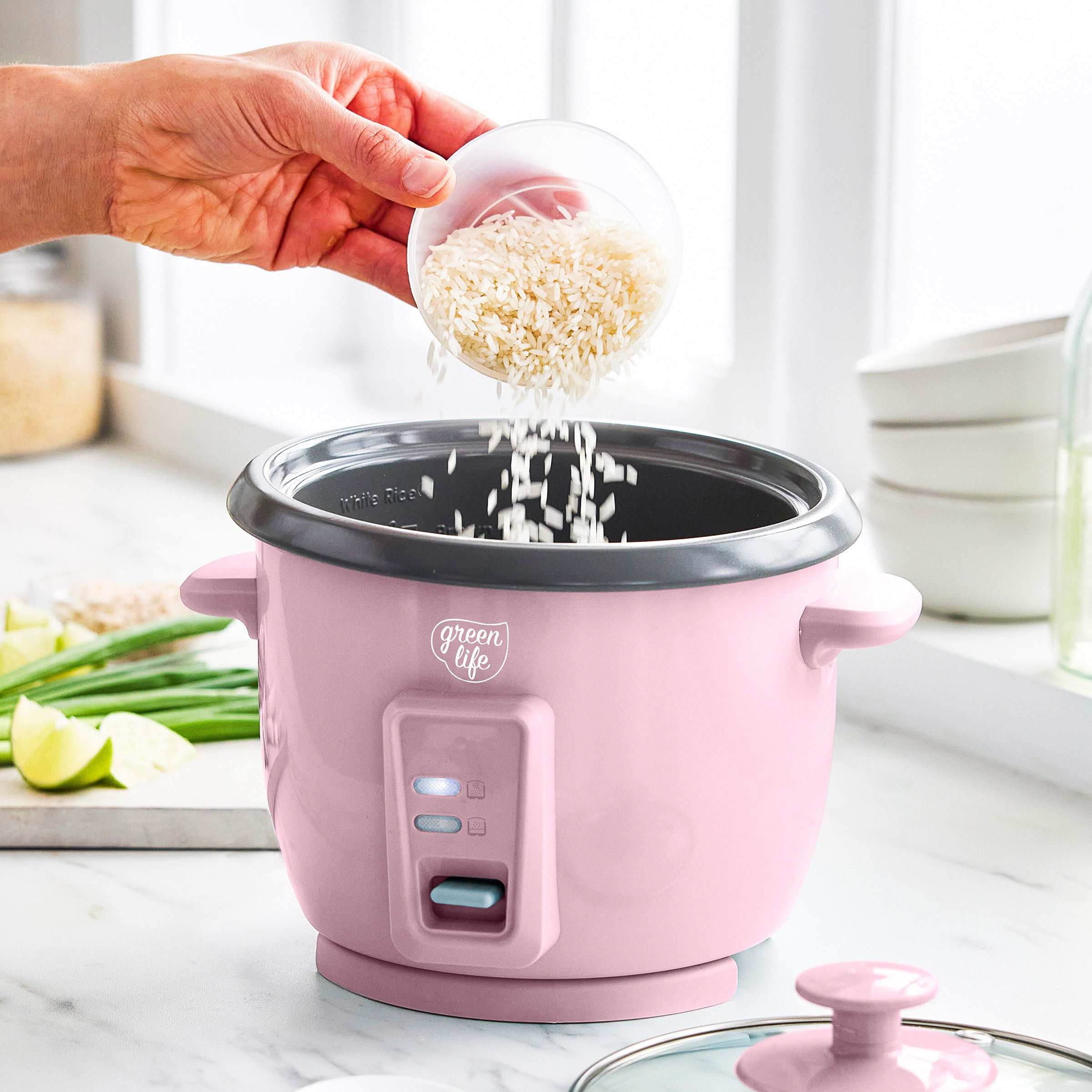 Pink rice cooker with a hand pouring rice into it on a kitchen counter.