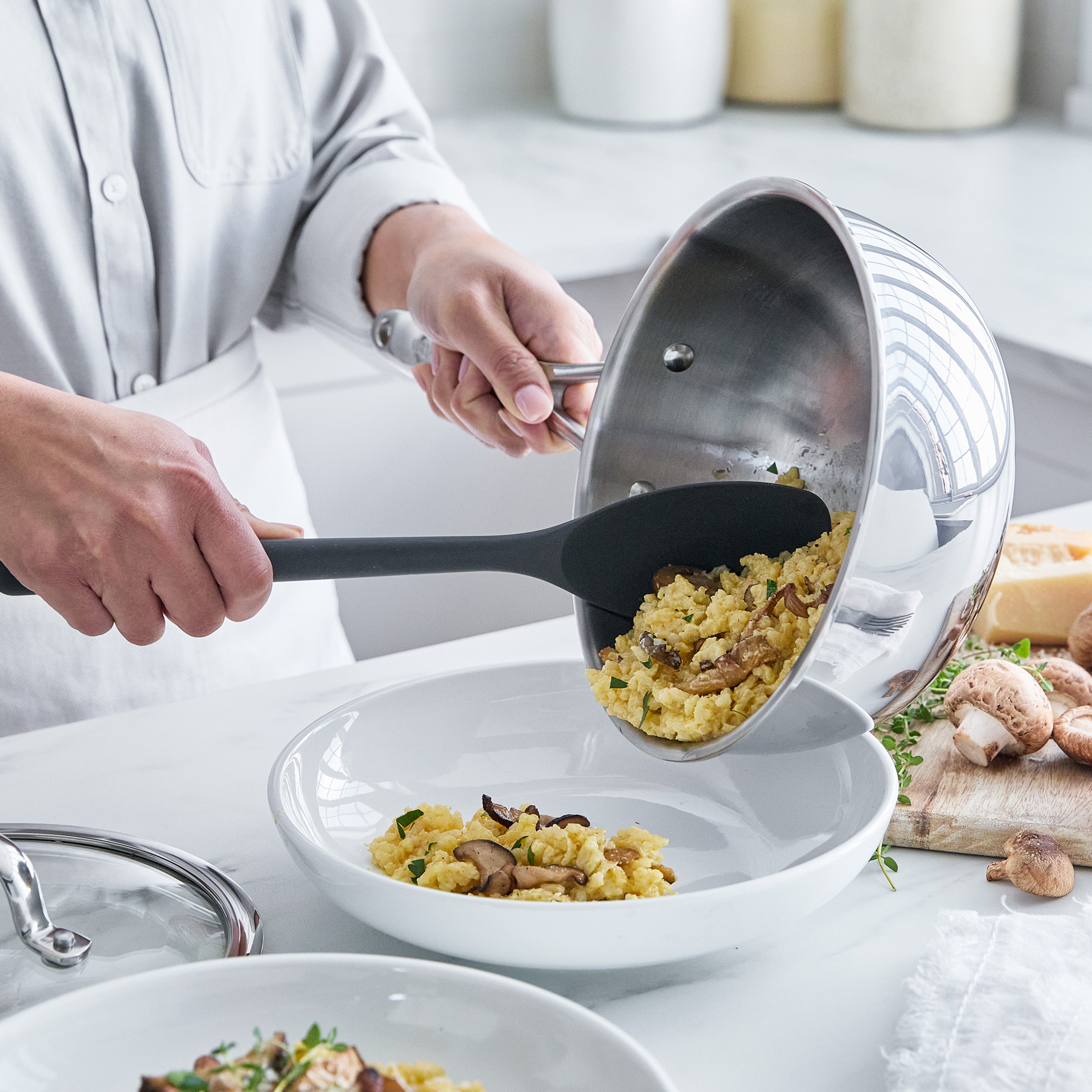 Person serving food from a stainless steel chef's pan into a white bowl with a black spoon.