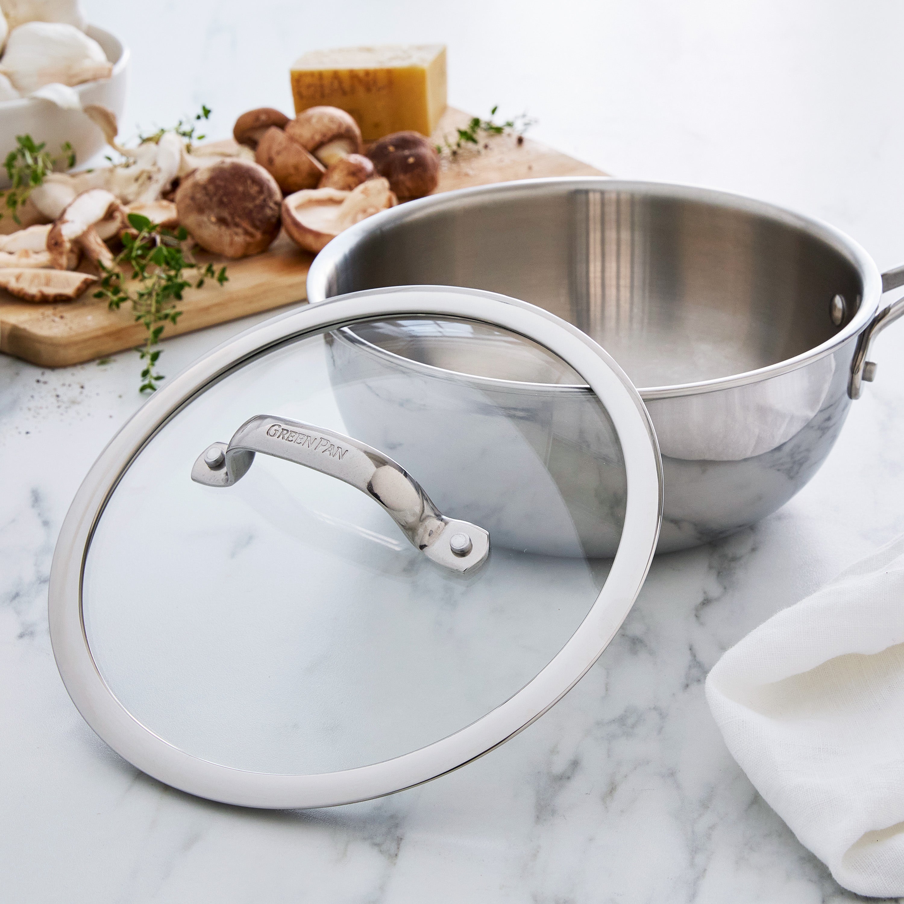 Stainless steel chef's pan with glass lid on a marble surface with mushrooms and herbs in the background.