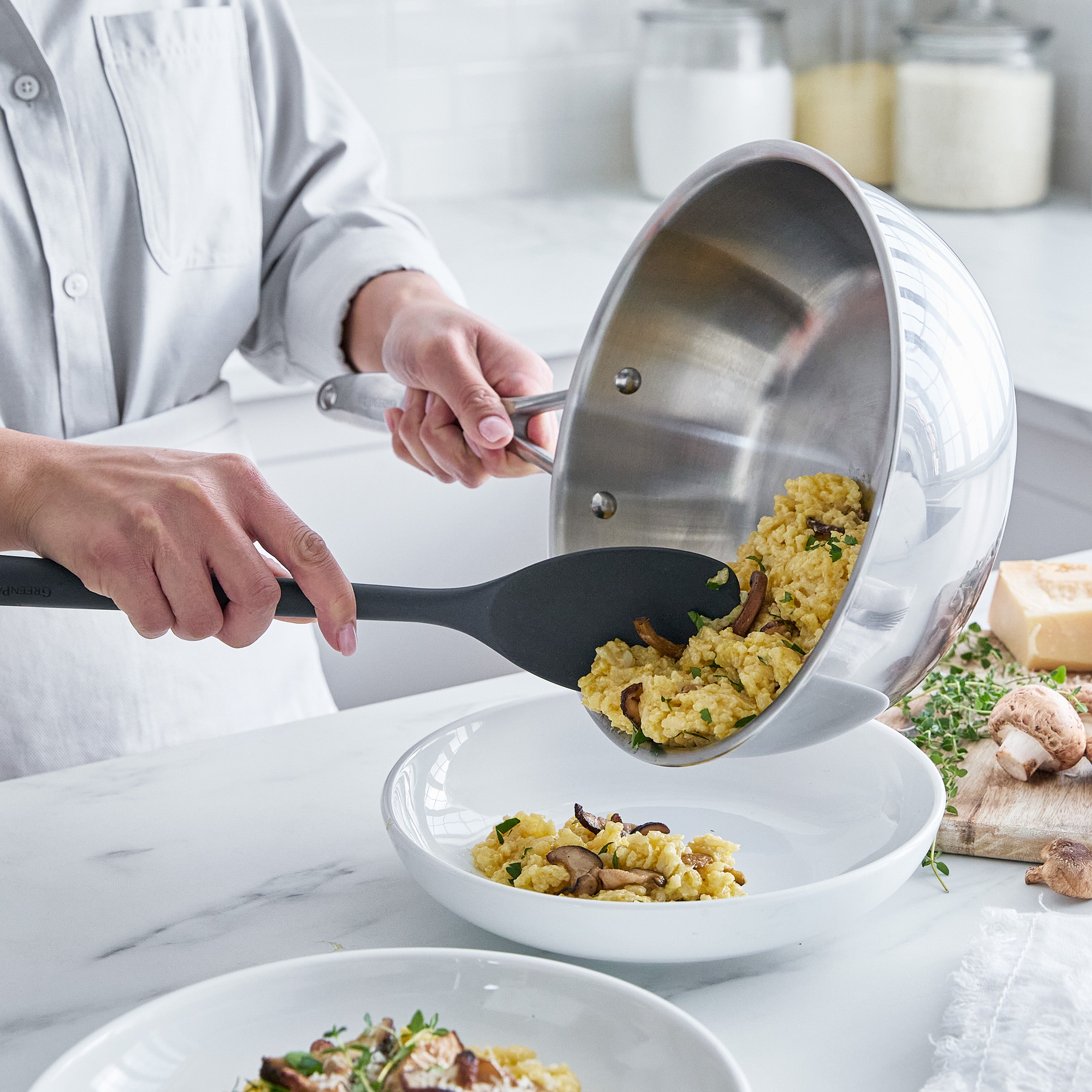 Person serving pasta with mushrooms from a chef's pan into a white bowl on a marble countertop.