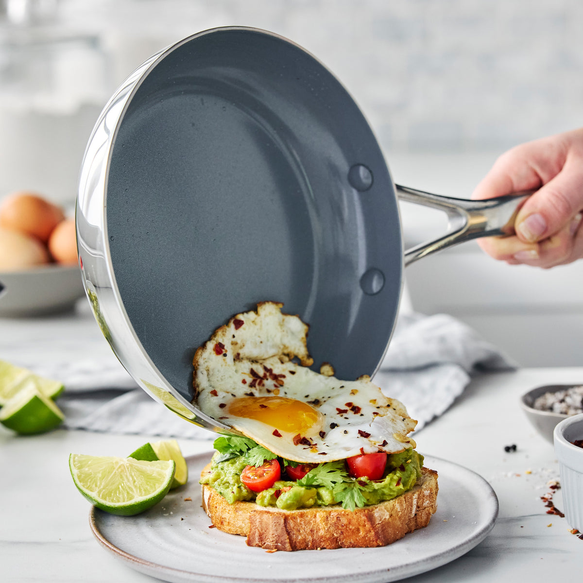 Fried egg sliding from a stainless steel Valencia Pro 9-piece cookware skillet onto avocado toast with cherry tomatoes.