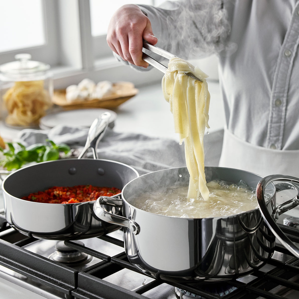 Tongs lifting steaming pasta from a Valencia Pro stainless steel 9-piece cookware pot next to a pan of tomatoes.