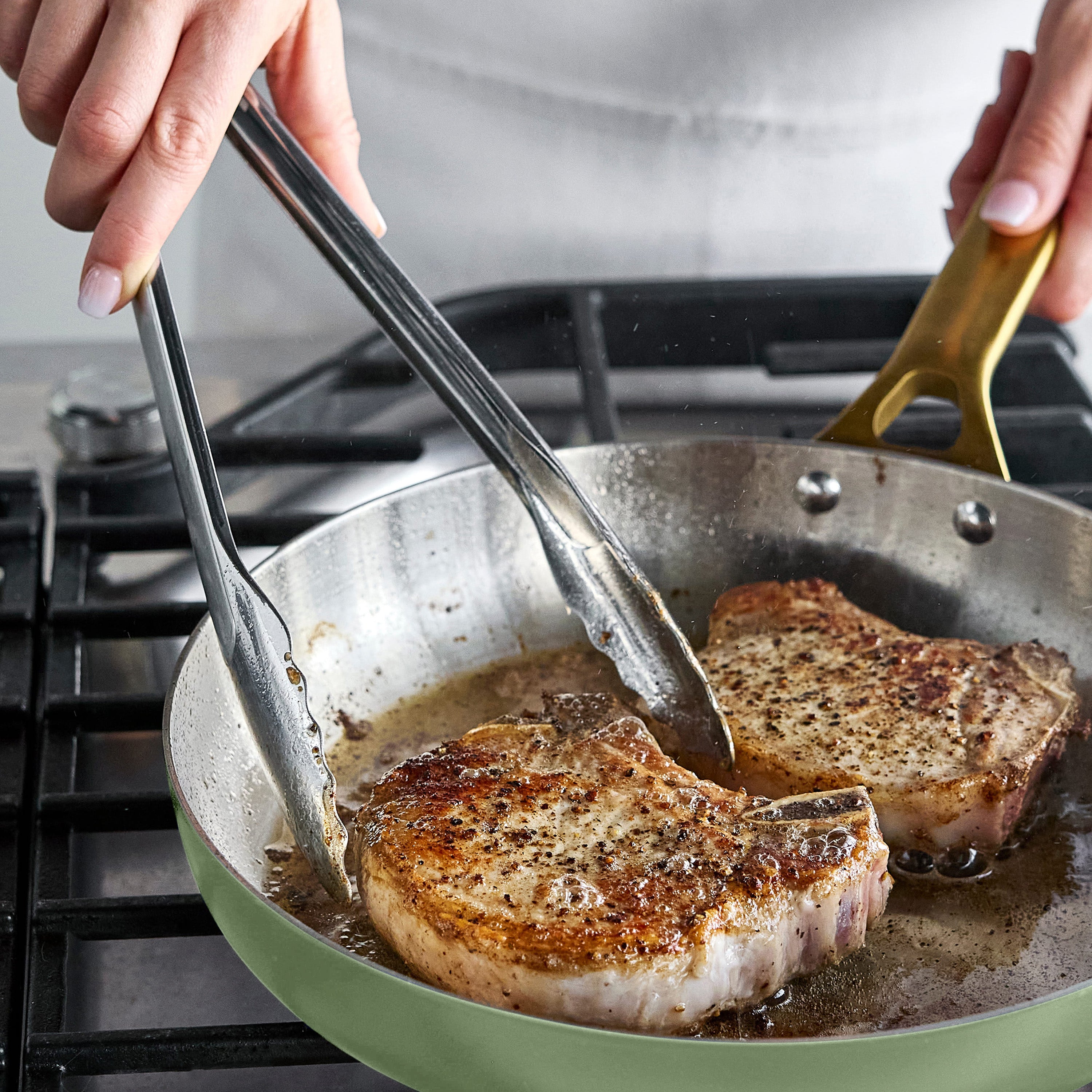 Person cooking pork chops in a pan with tongs on a stovetop.