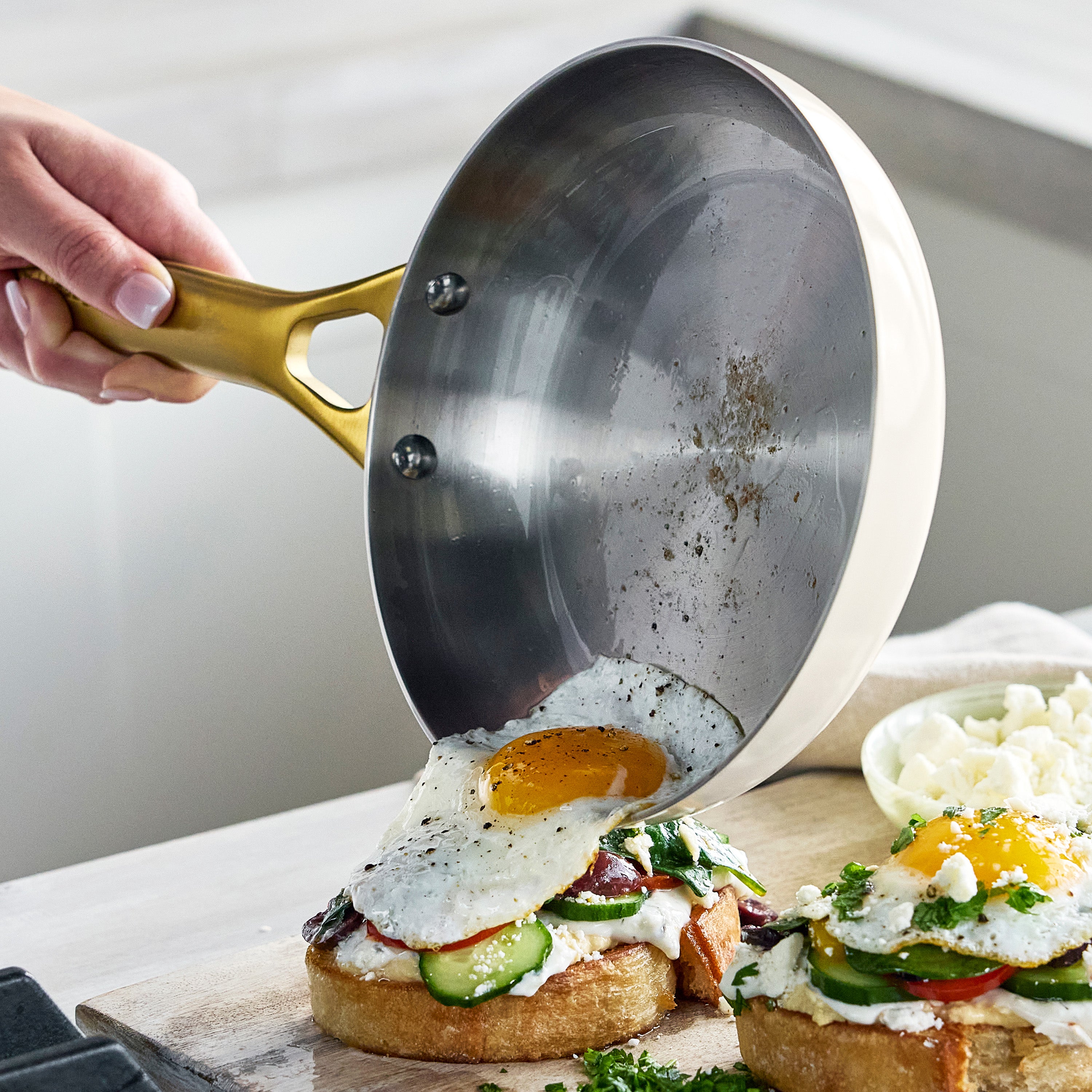 Person using a frying pan to place an egg onto a piece of toast with vegetables on a wooden cutting board.