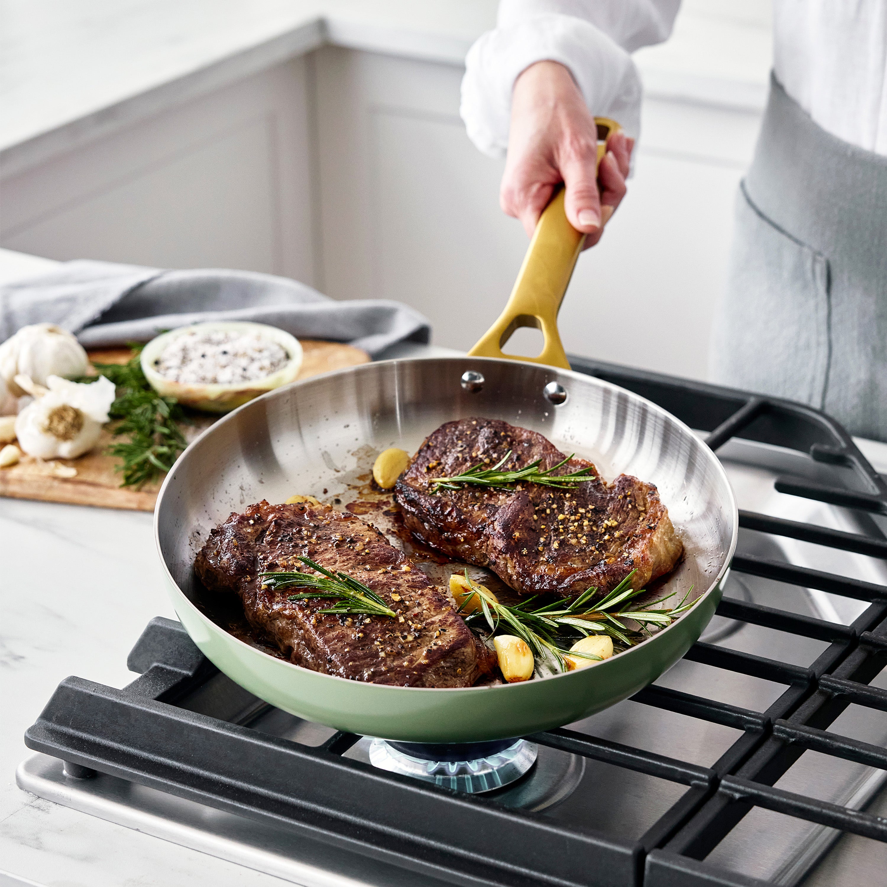 Steaks being cooked in a green frying pan on a stove with a person holding the handle.