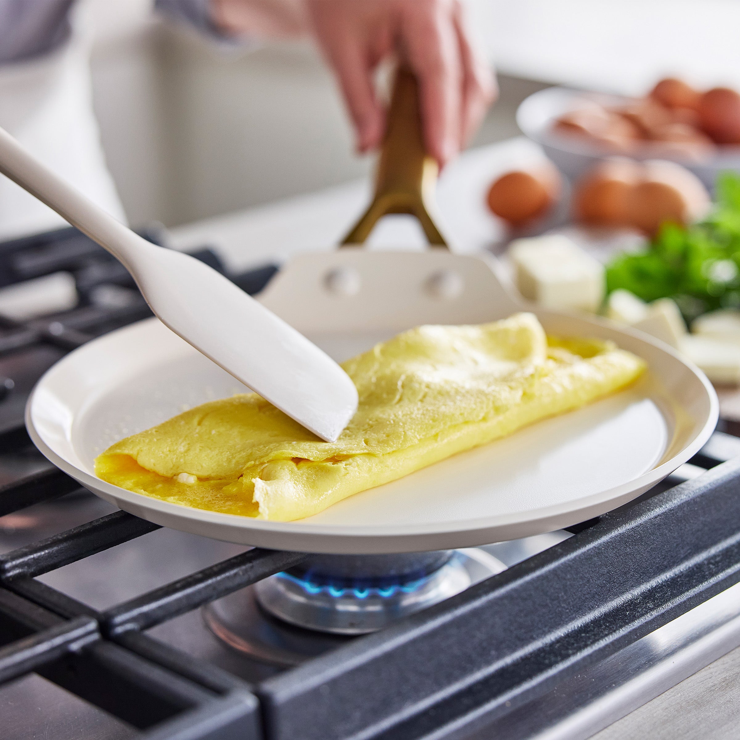 Omelette being folded on a crepe pan over a stove with eggs and herbs in the background