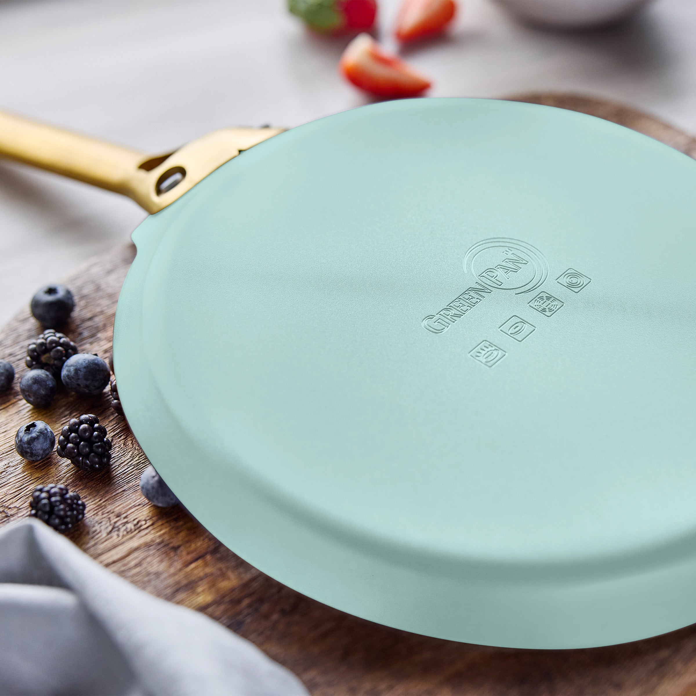 Light blue crepe pan with gold handle on a wooden surface with berries and strawberries.