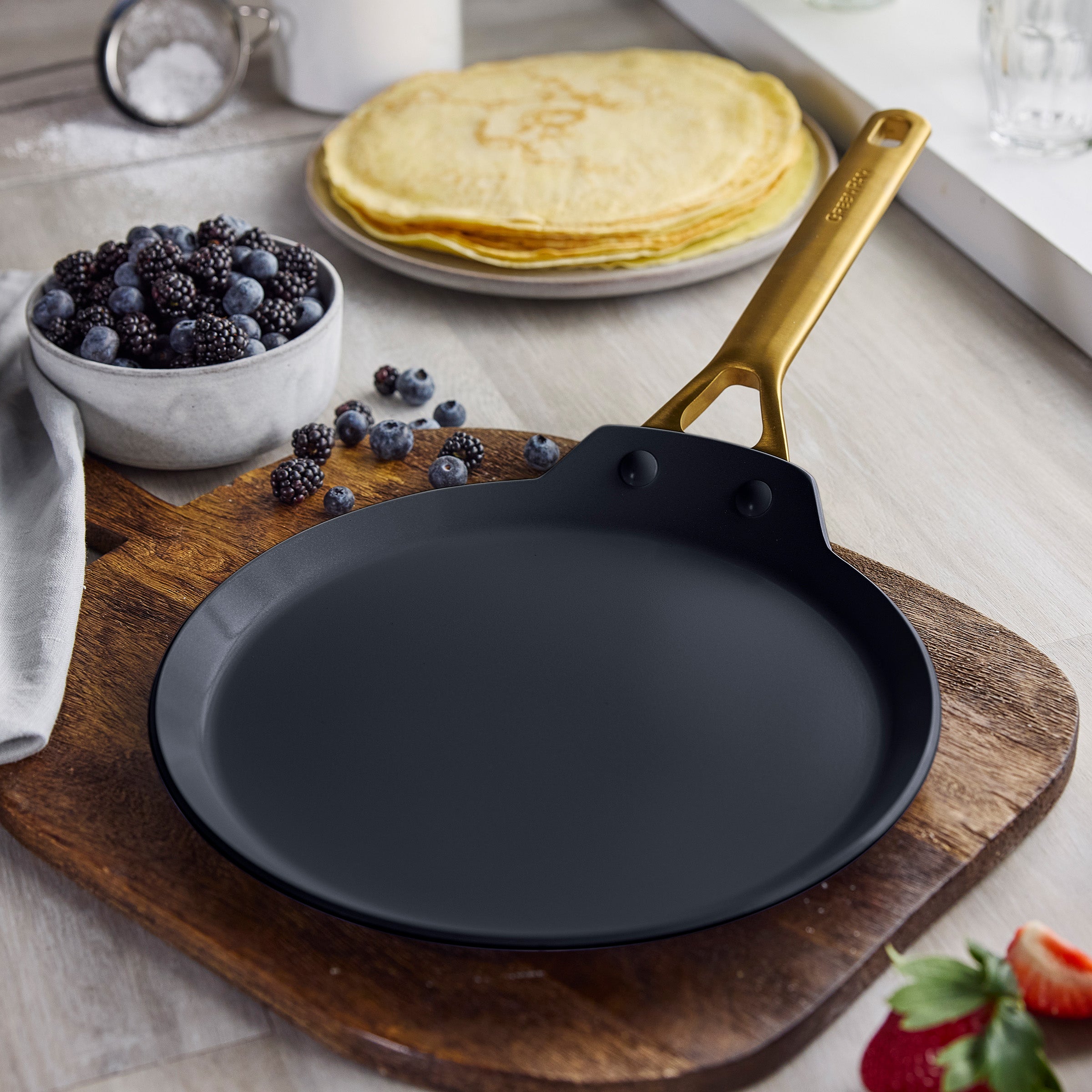 Black crepe pan on a wooden cutting board with crepes and berries in the background