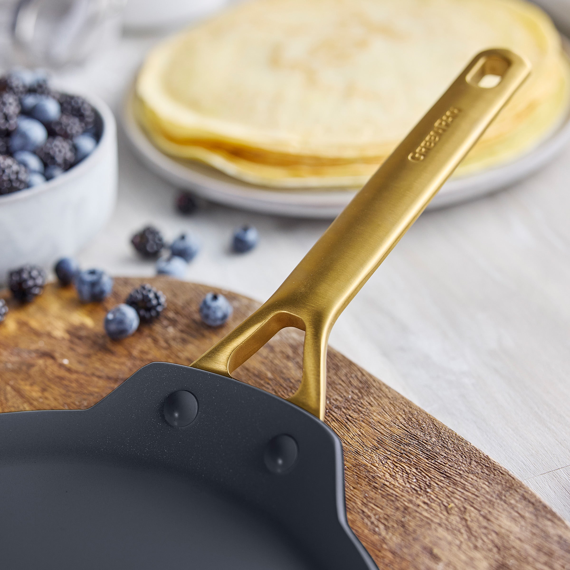 Black crepr pan with gold handle on a wooden cutting board, with berries and crepes in the background.