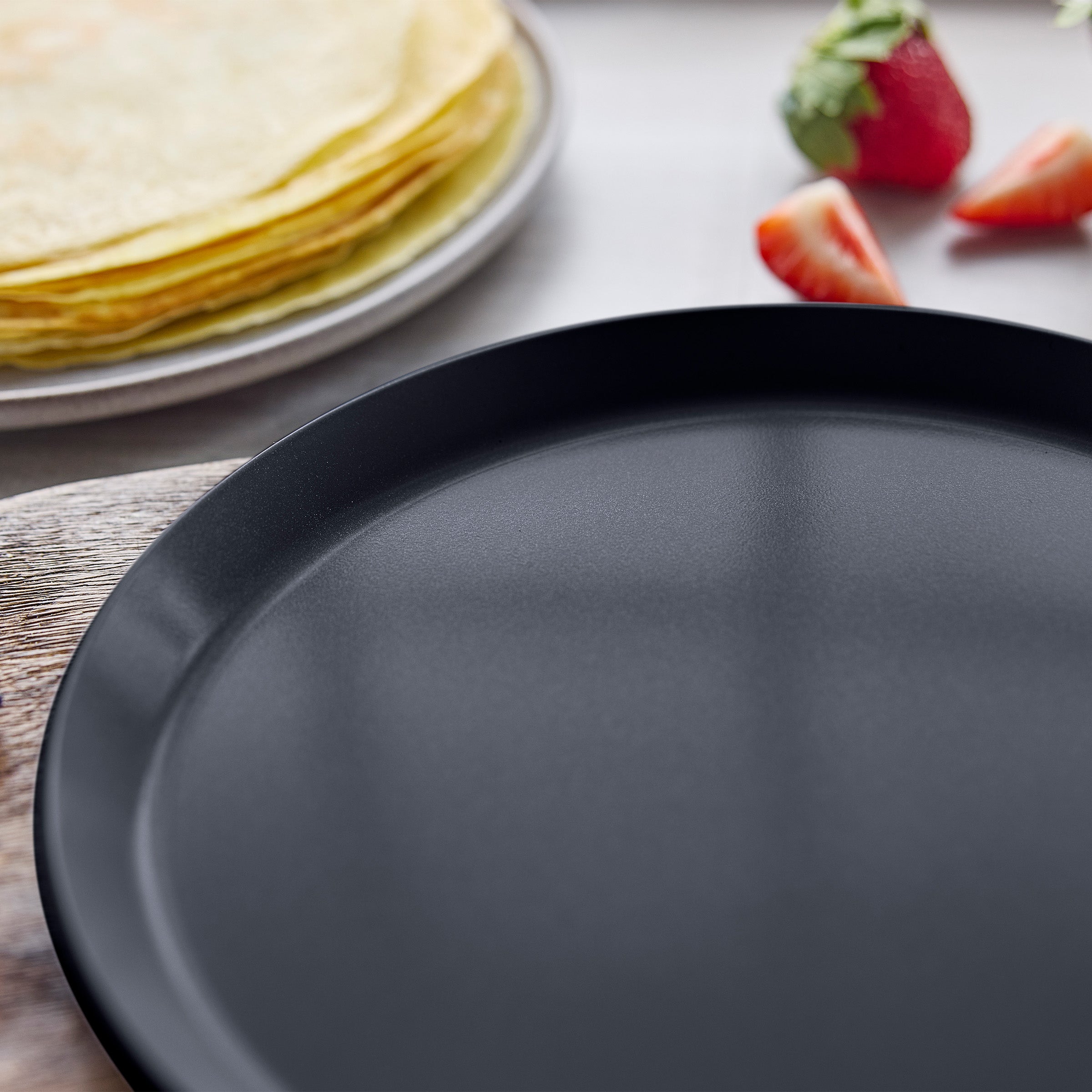 Black crepe pan on a wooden surface with crepes and strawberries in the background