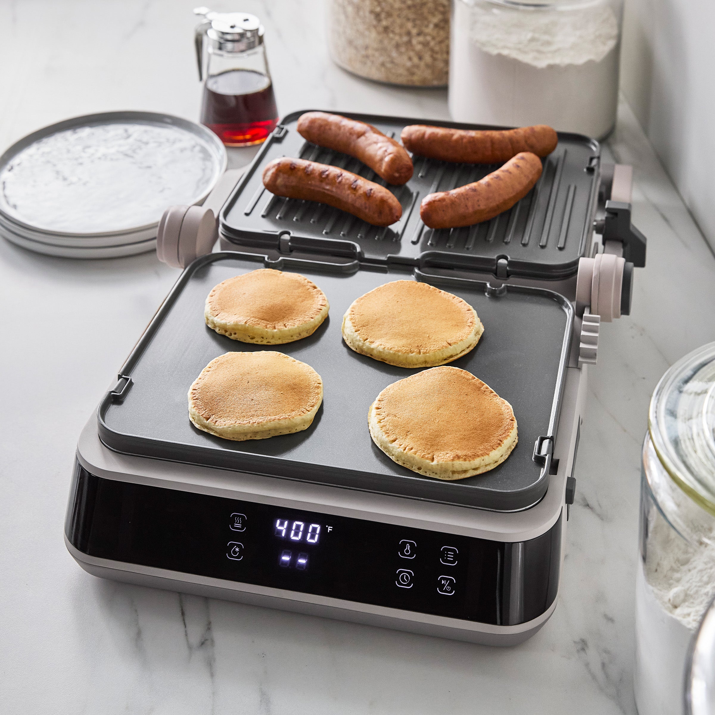 Ceramic nonstick grill and griddle with digital display, pancakes and sausages cooking, syrup jars on a marble countertop.