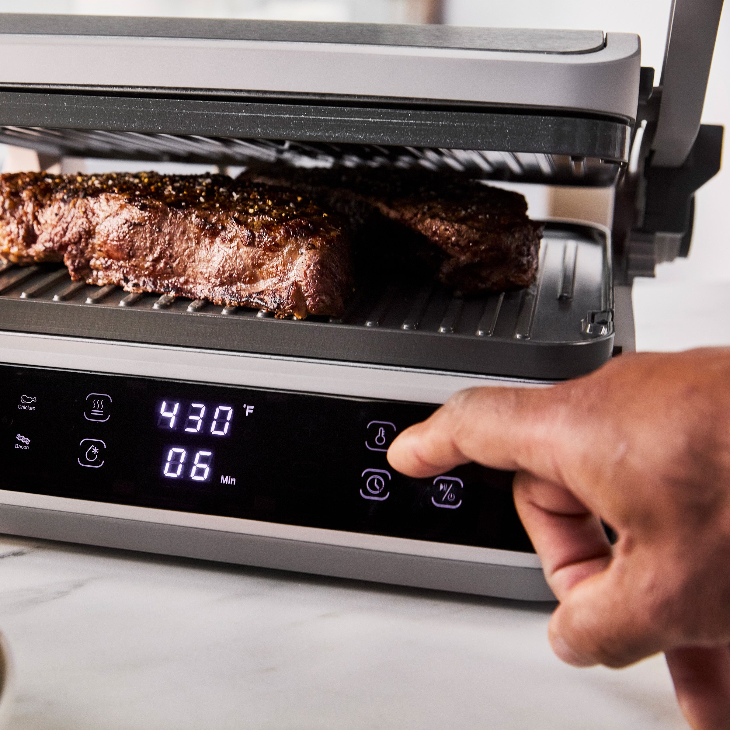 Hand pressing a button on the Advanced Grill and Griddle set to 430°F, grilling steaks on a ceramic plate.