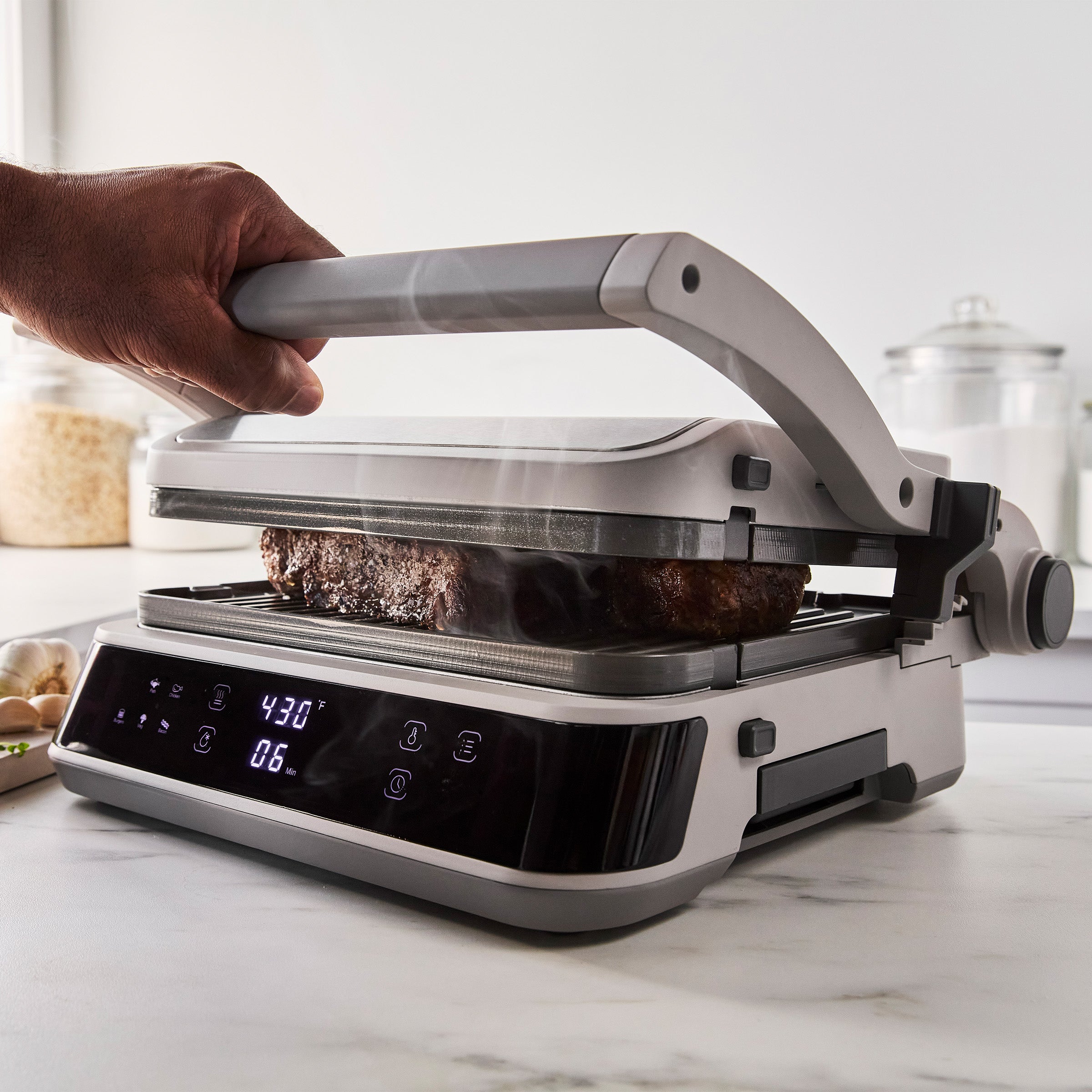 Hand pressing a steak on the Advanced Grill and Griddle | Clay, steam rising from the meat on a white countertop.