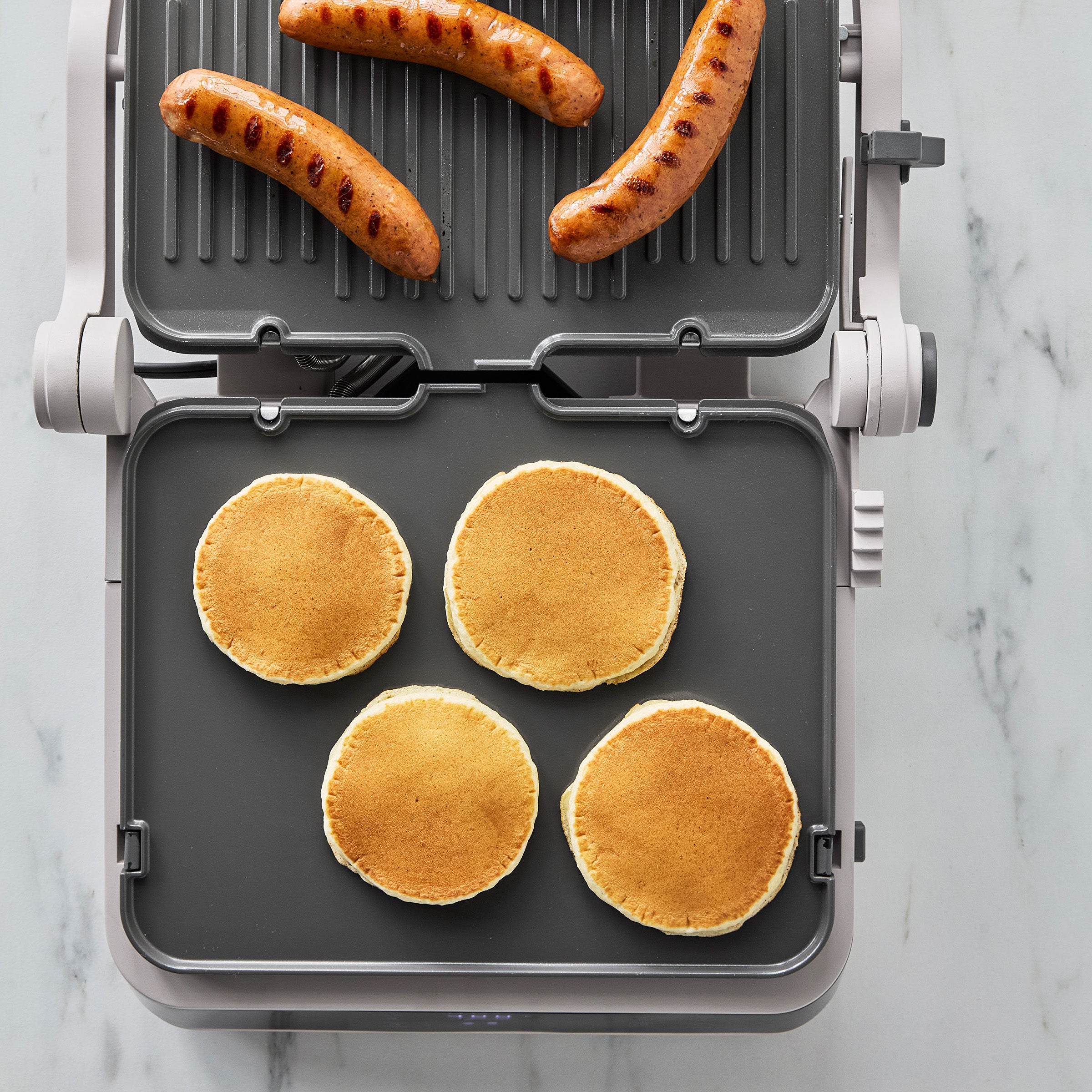 Four pancakes and three sausages cooking on the clay Advanced Grill and Griddle, viewed from above.