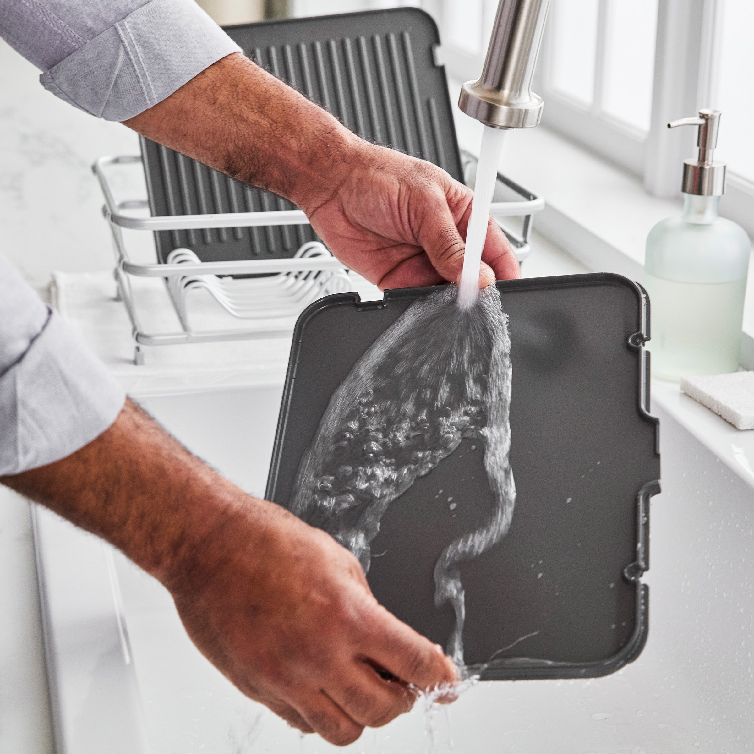 Clay grill and griddle being washed under running water next to a dish rack and soap dispenser in a kitchen sink.