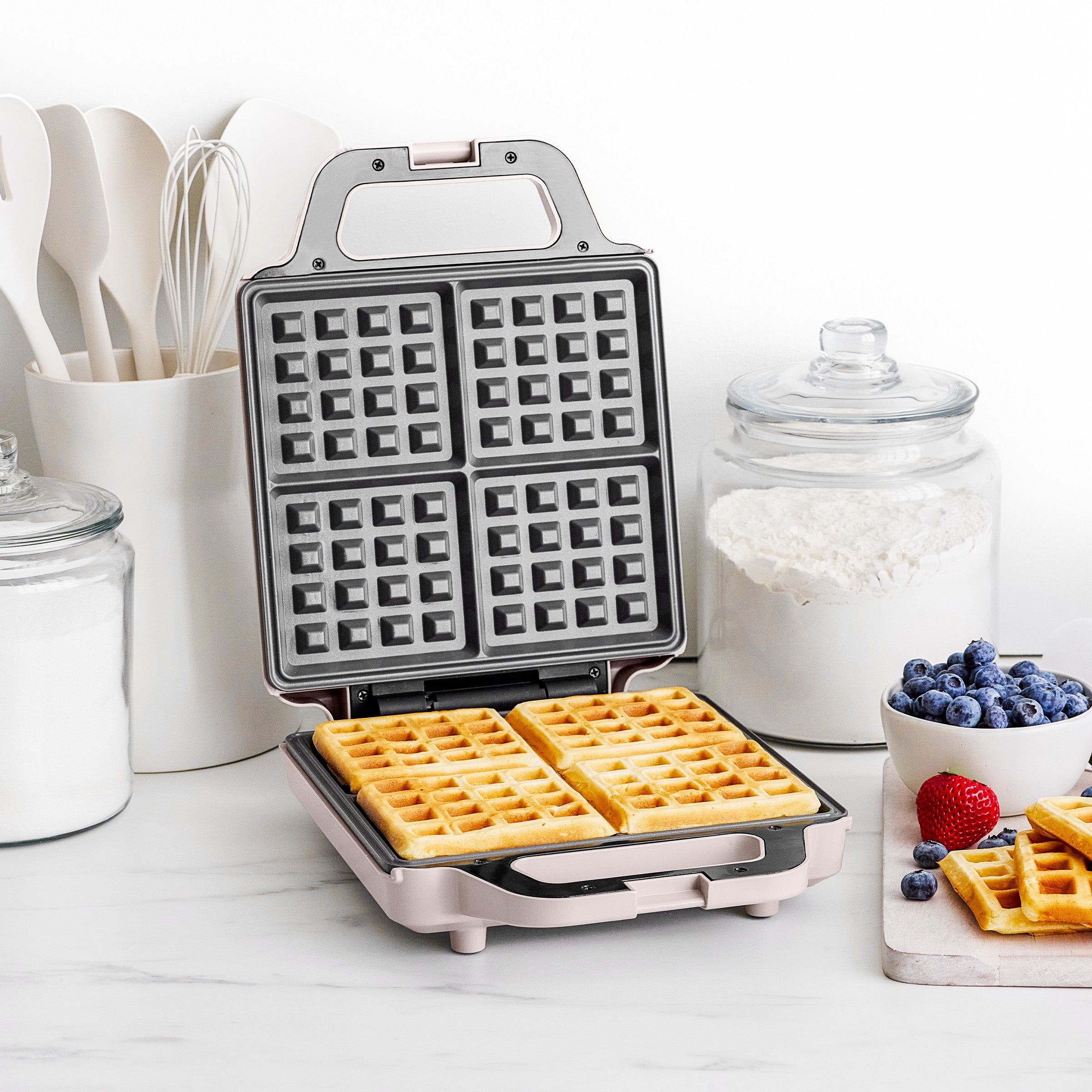 Waffle maker with waffles on a kitchen counter with ingredients and utensils.