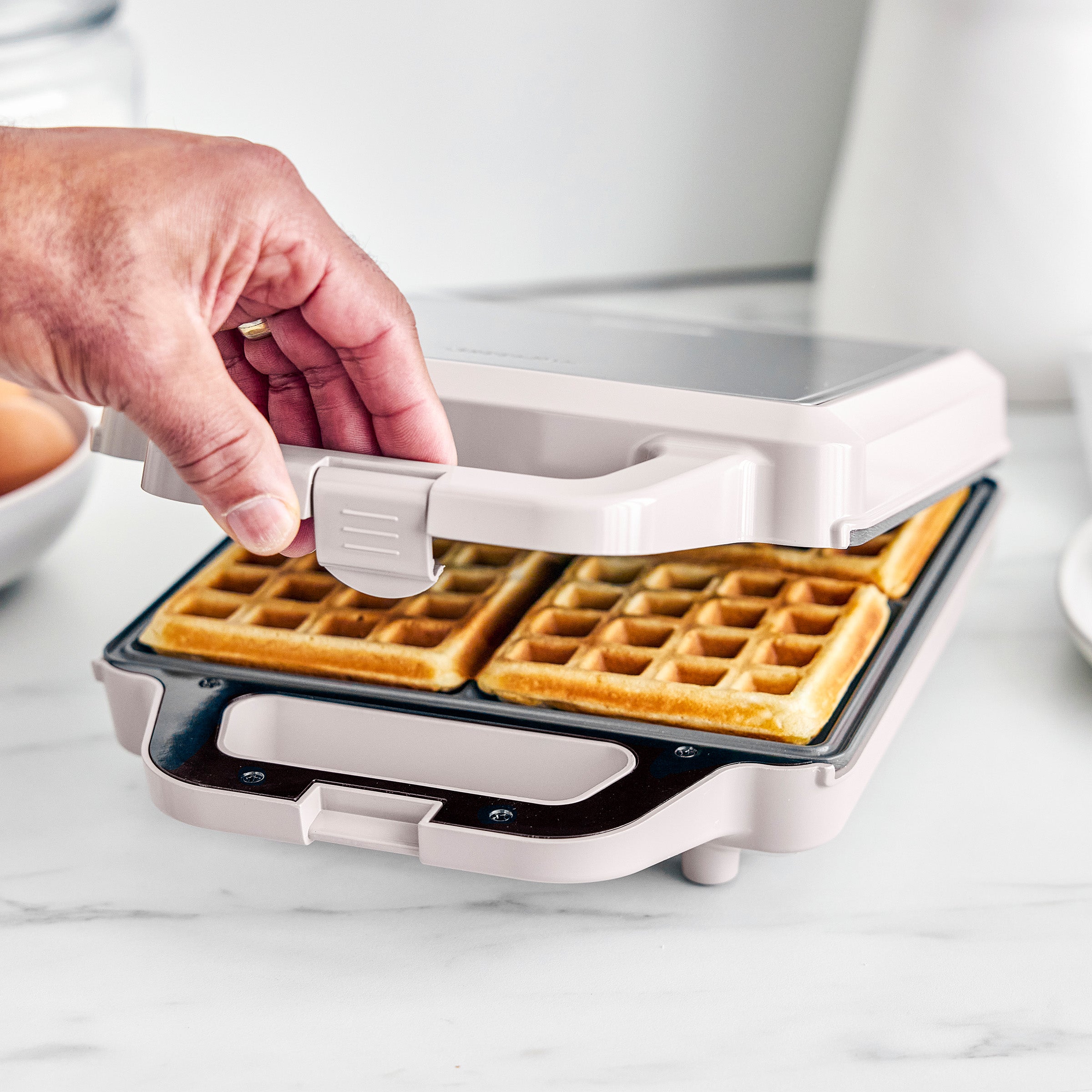 Person lifting a waffle from a white waffle maker on a light surface.