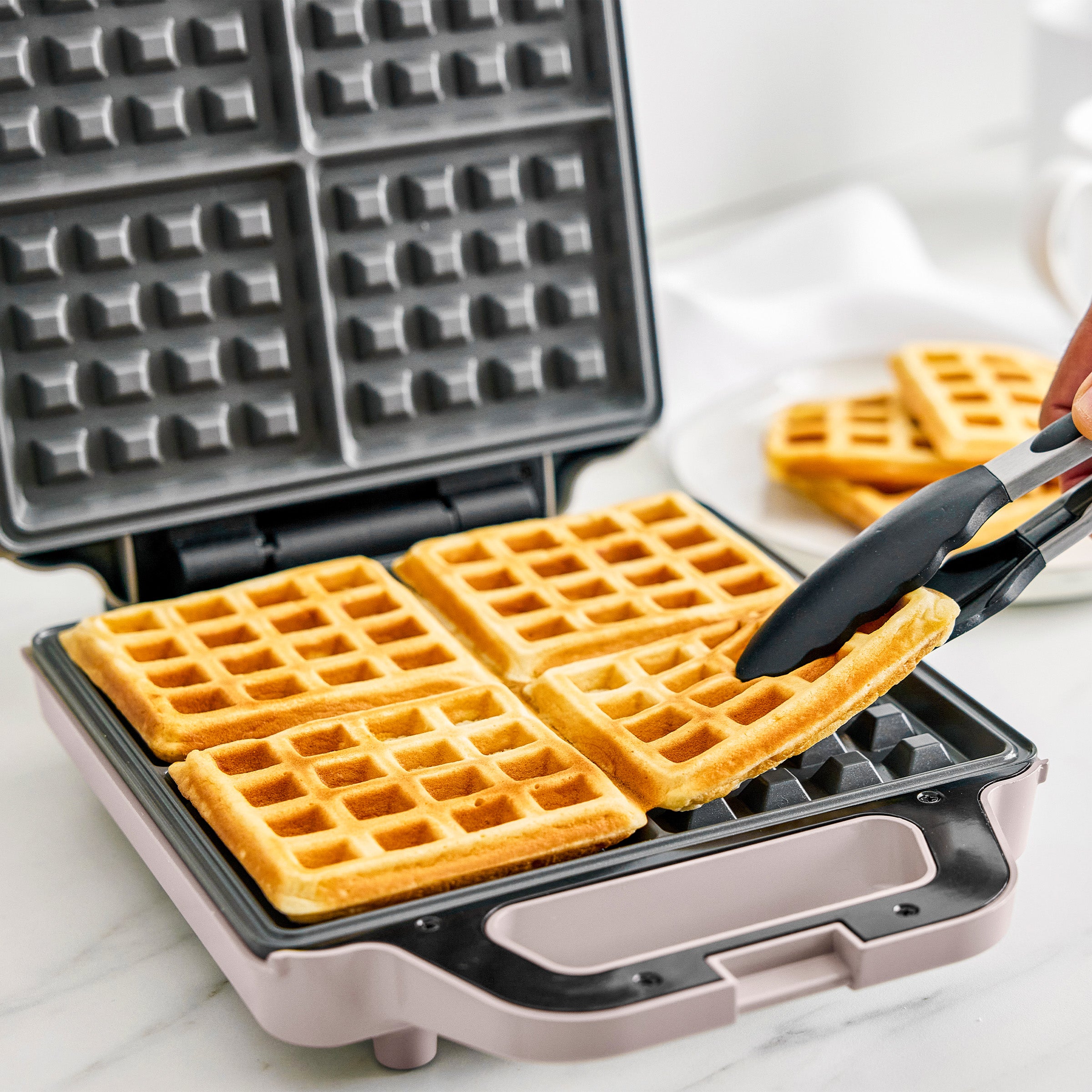 Waffle maker with freshly made waffles and a hand using tongs to lift one.