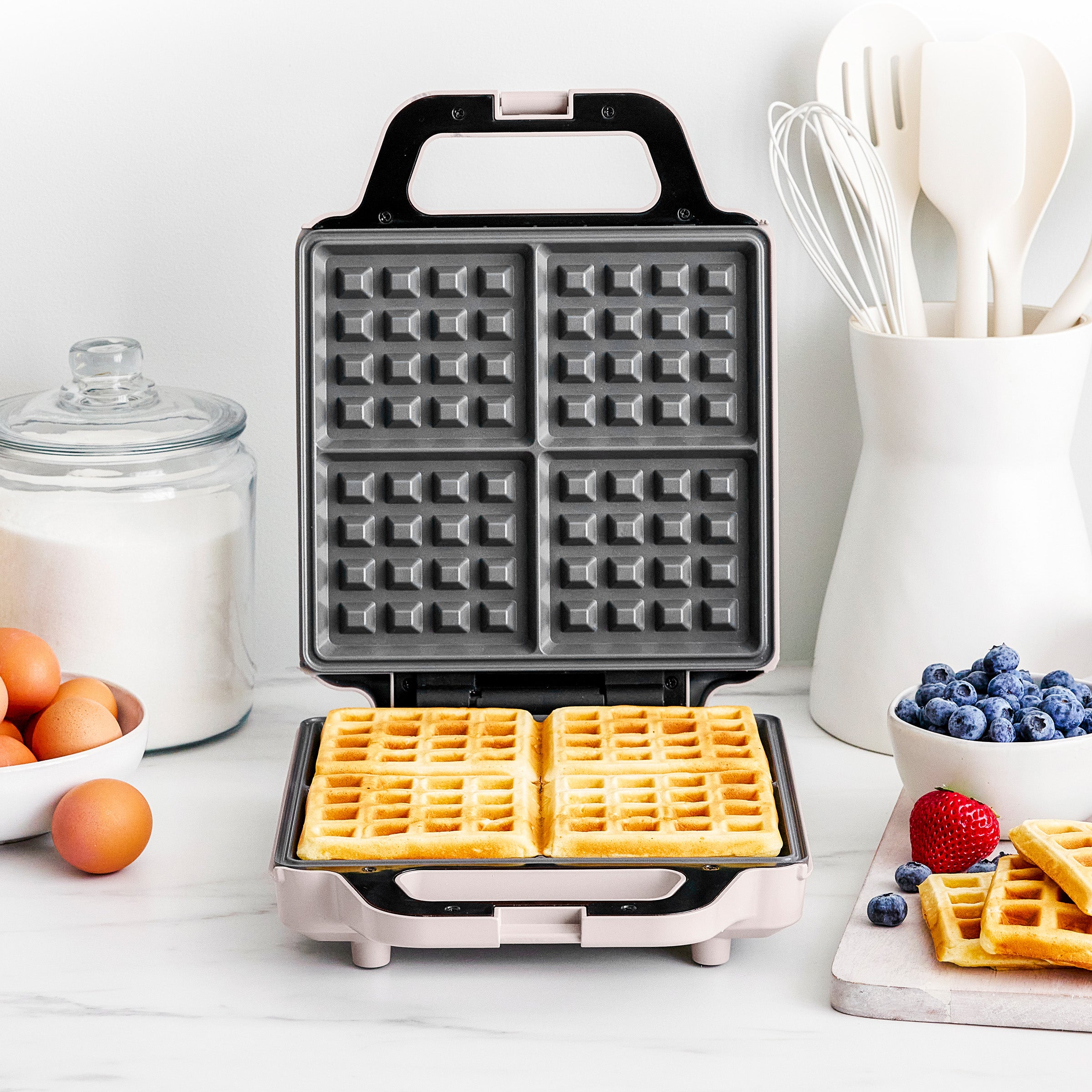 Waffle maker with waffles on a kitchen counter with ingredients and utensils.