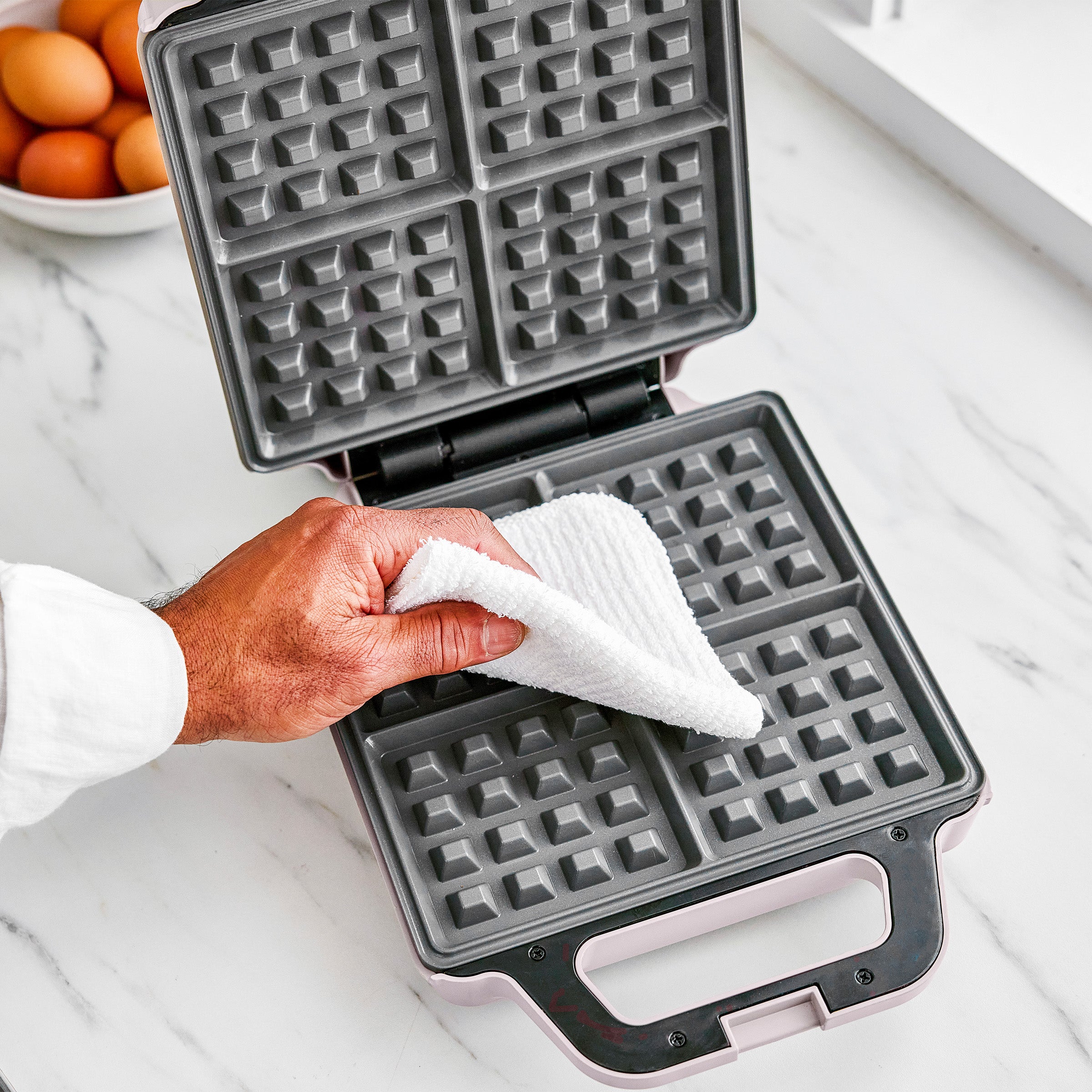 Person cleaning a waffle maker with a white cloth on a marble countertop.