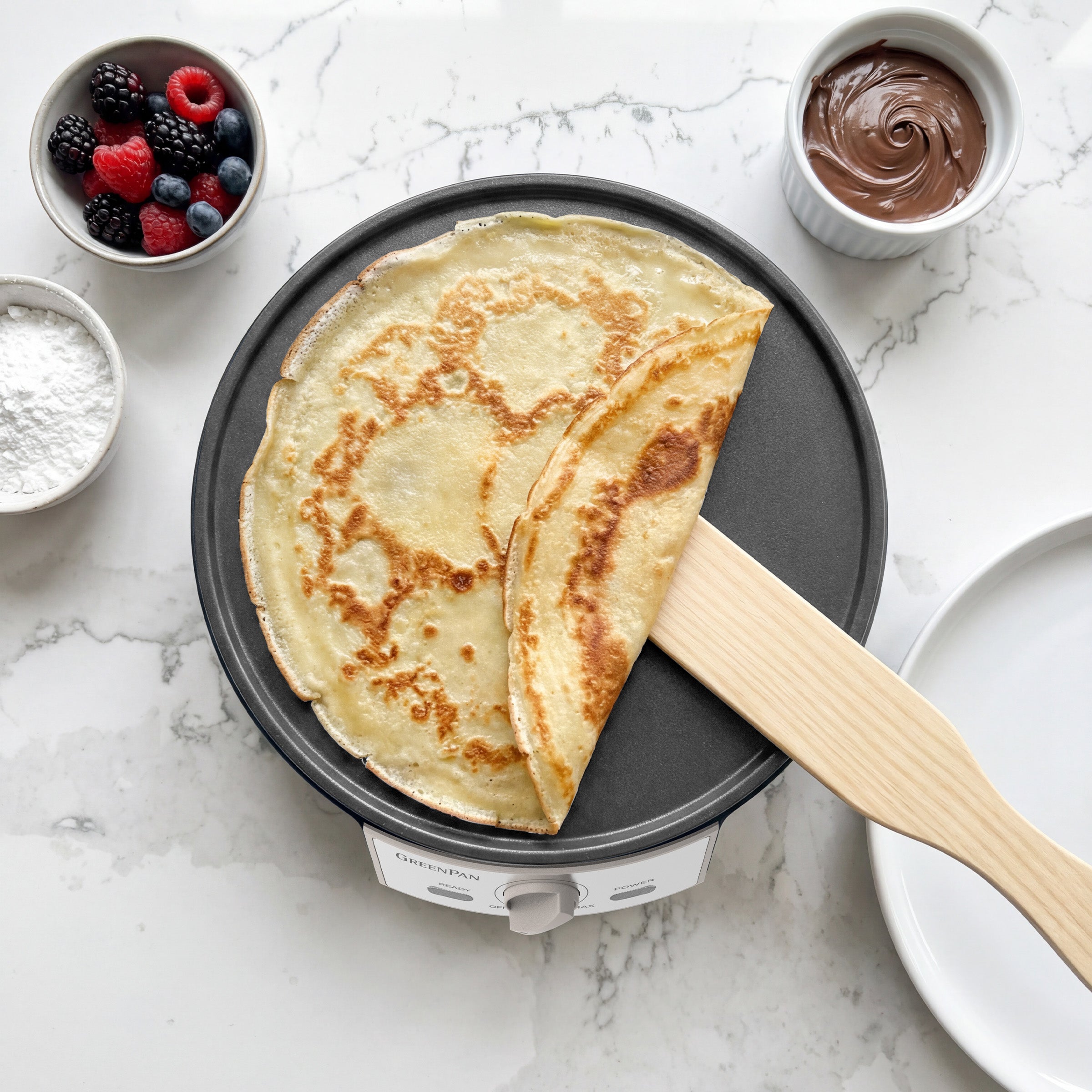 Crepe on a crepe maker with berries and Nutella nearby