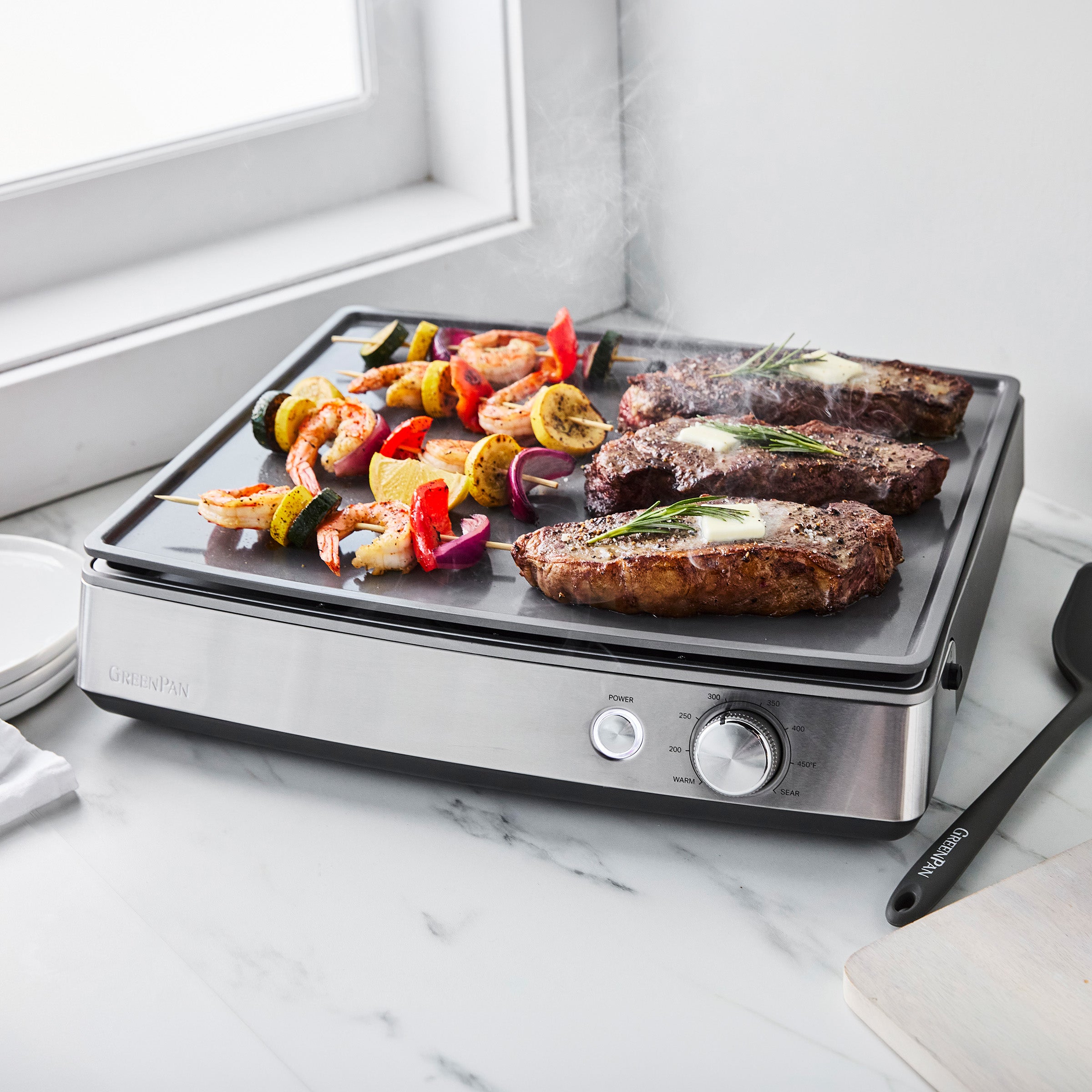 Three steaks and assorted vegetables cooking on a stainless steel countertop griddle near a kitchen window.