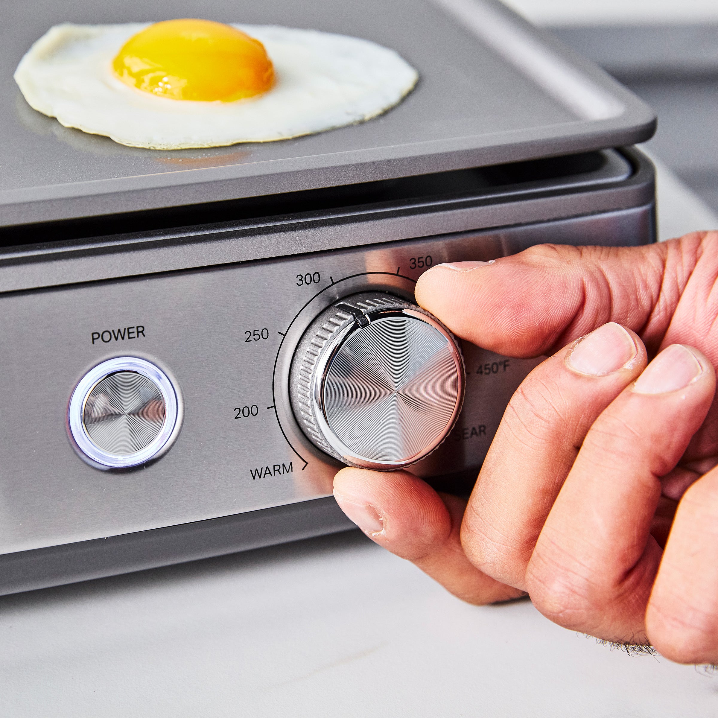 Hand adjusting stainless steel countertop griddle with a fried egg cooking on the nonstick surface.