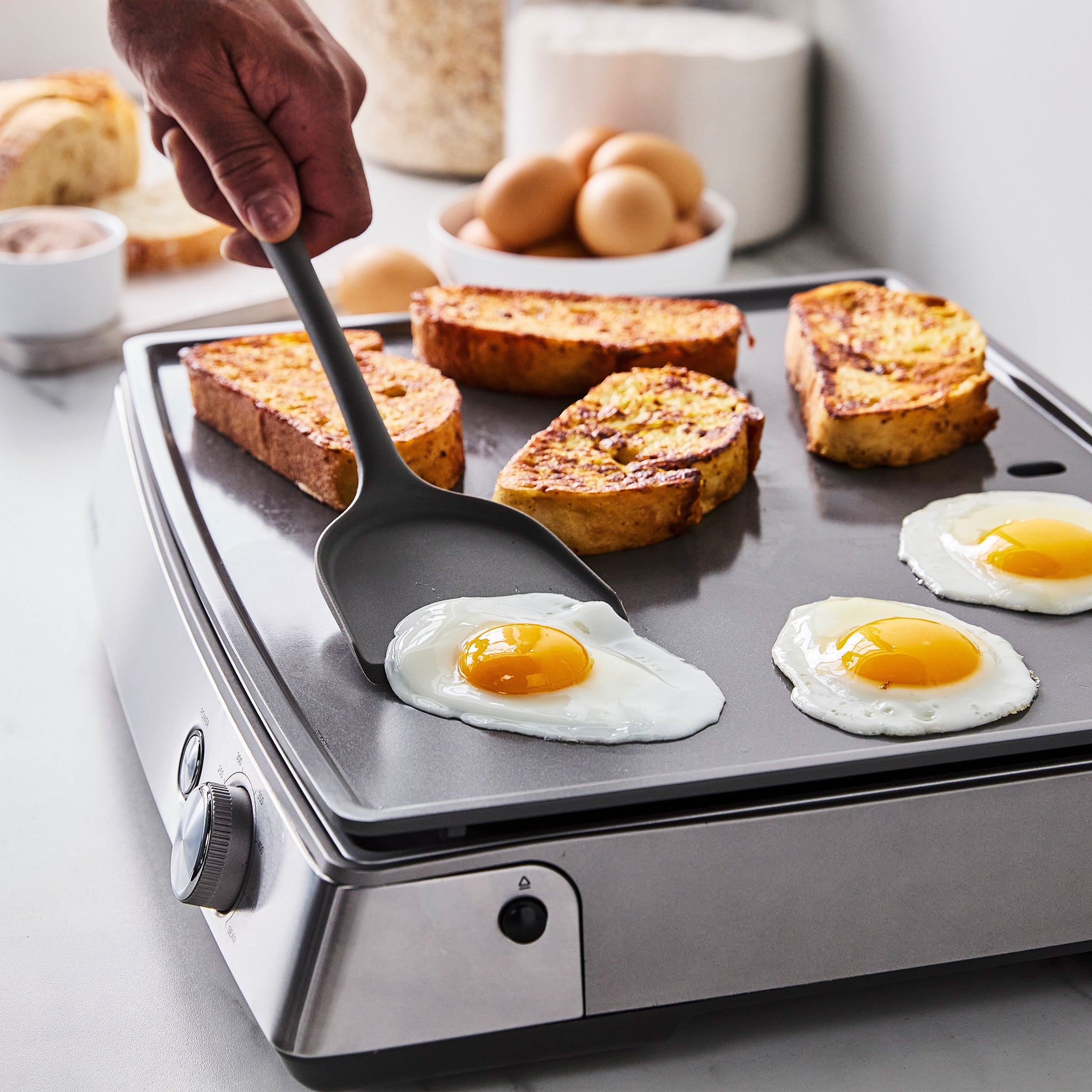 Hand flipping a fried egg on a flat countertop griddle, with eggs, French toast, and flour nearby.
