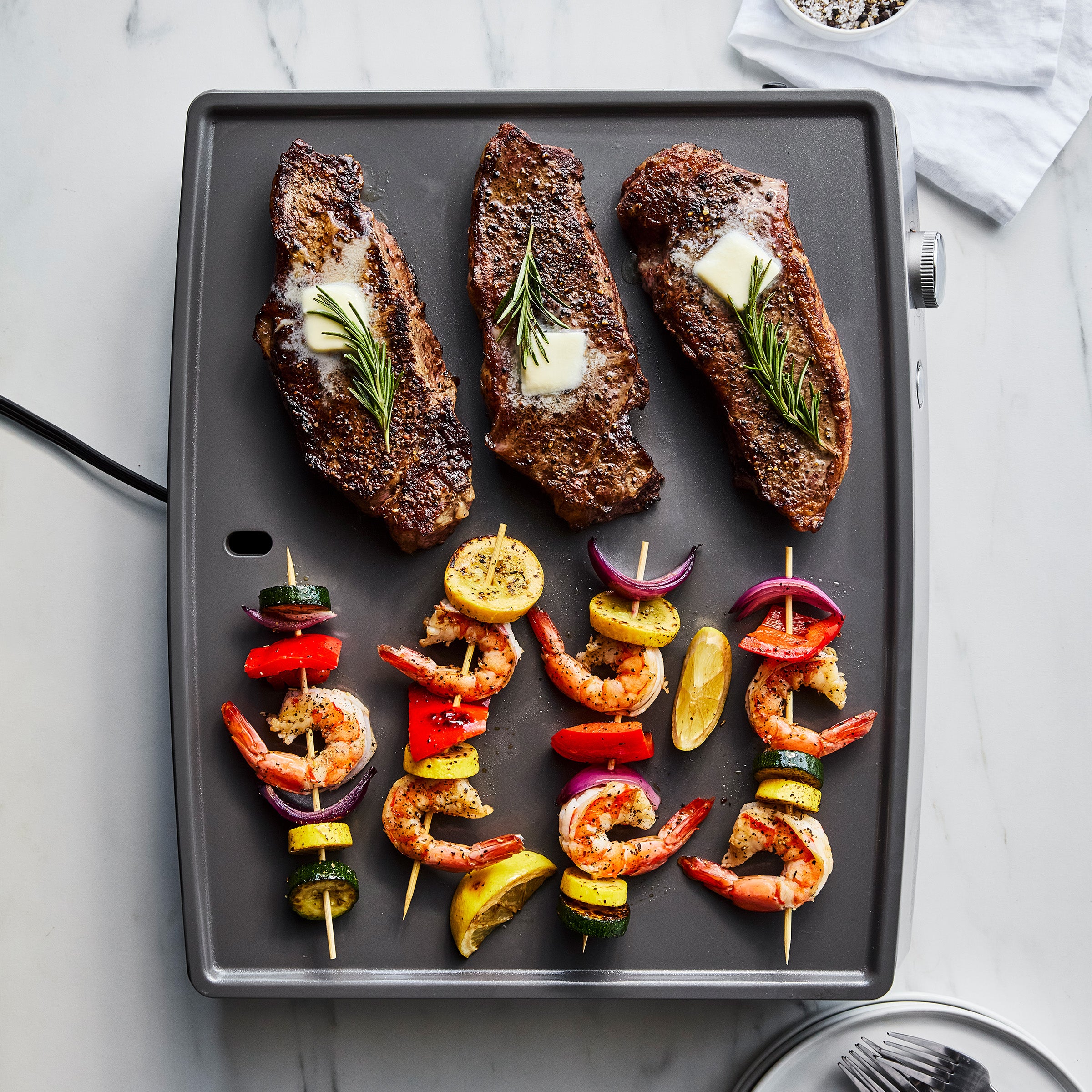 Three steaks and two shrimp-veggie skewers cooking on a stainless steel countertop griddle.