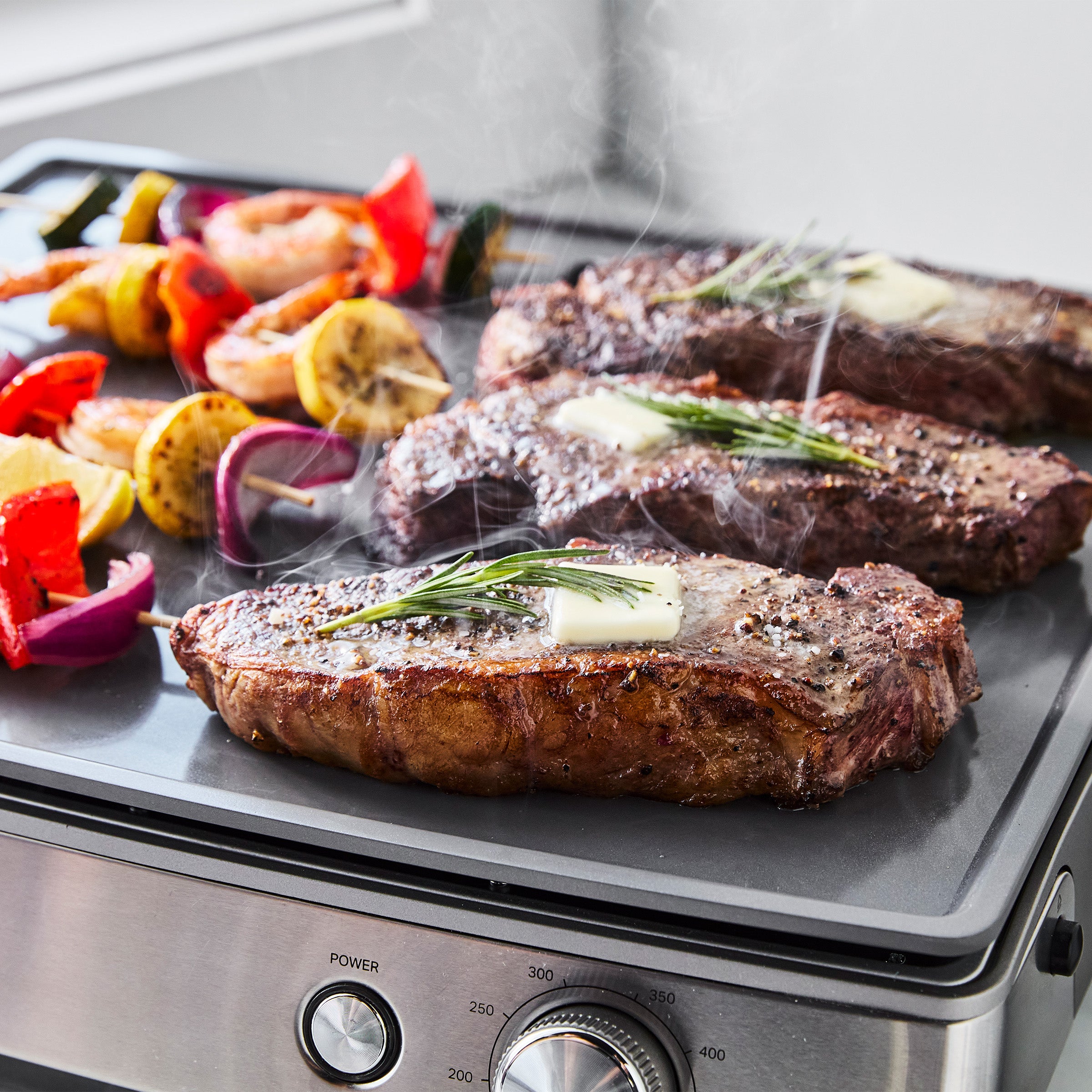 Steaks, butter, and rosemary cooking on a countertop griddle next to veggie-shrimp skewers with steam rising.