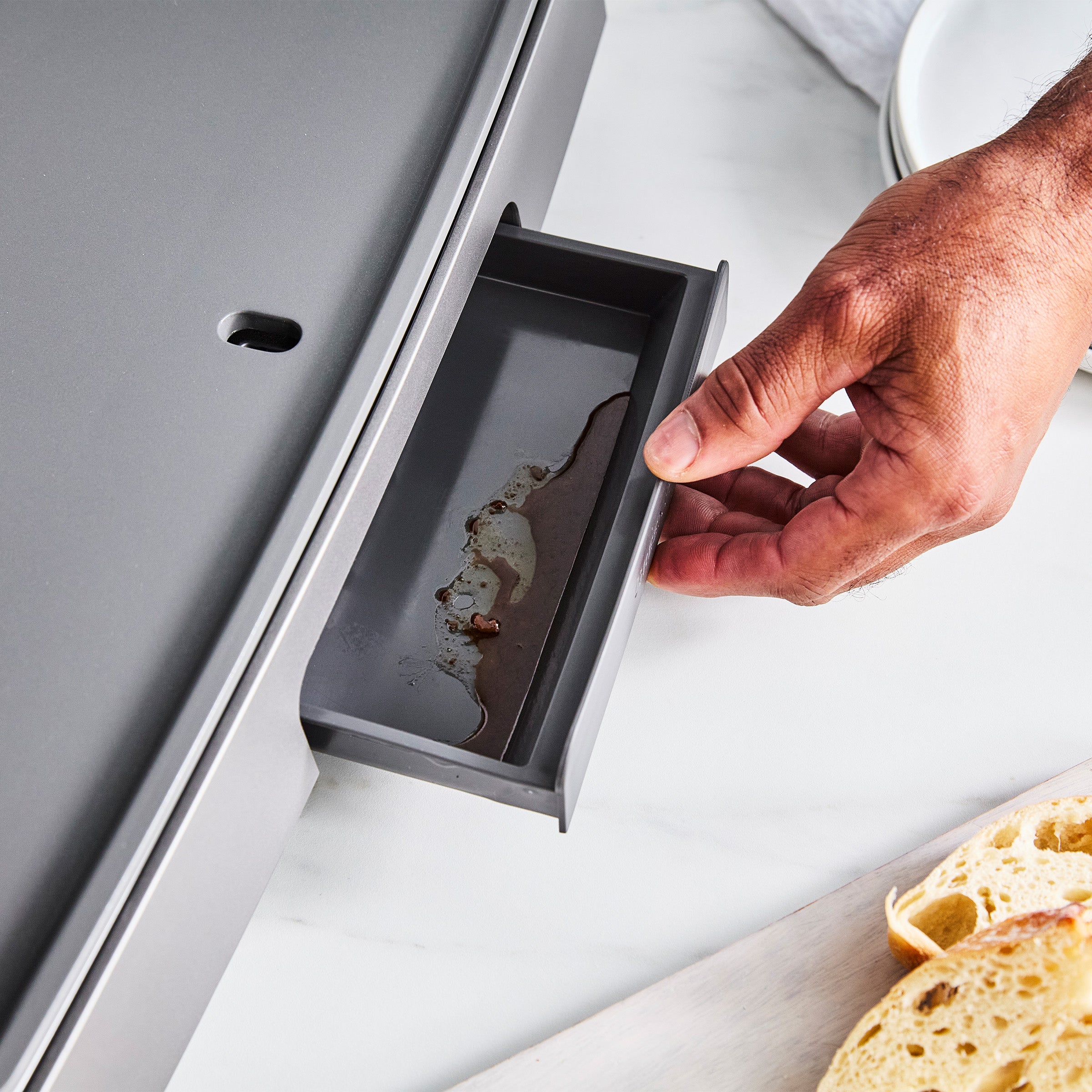 Hand pulling out grease tray from stainless steel countertop griddle; sliced bread on a white surface nearby.