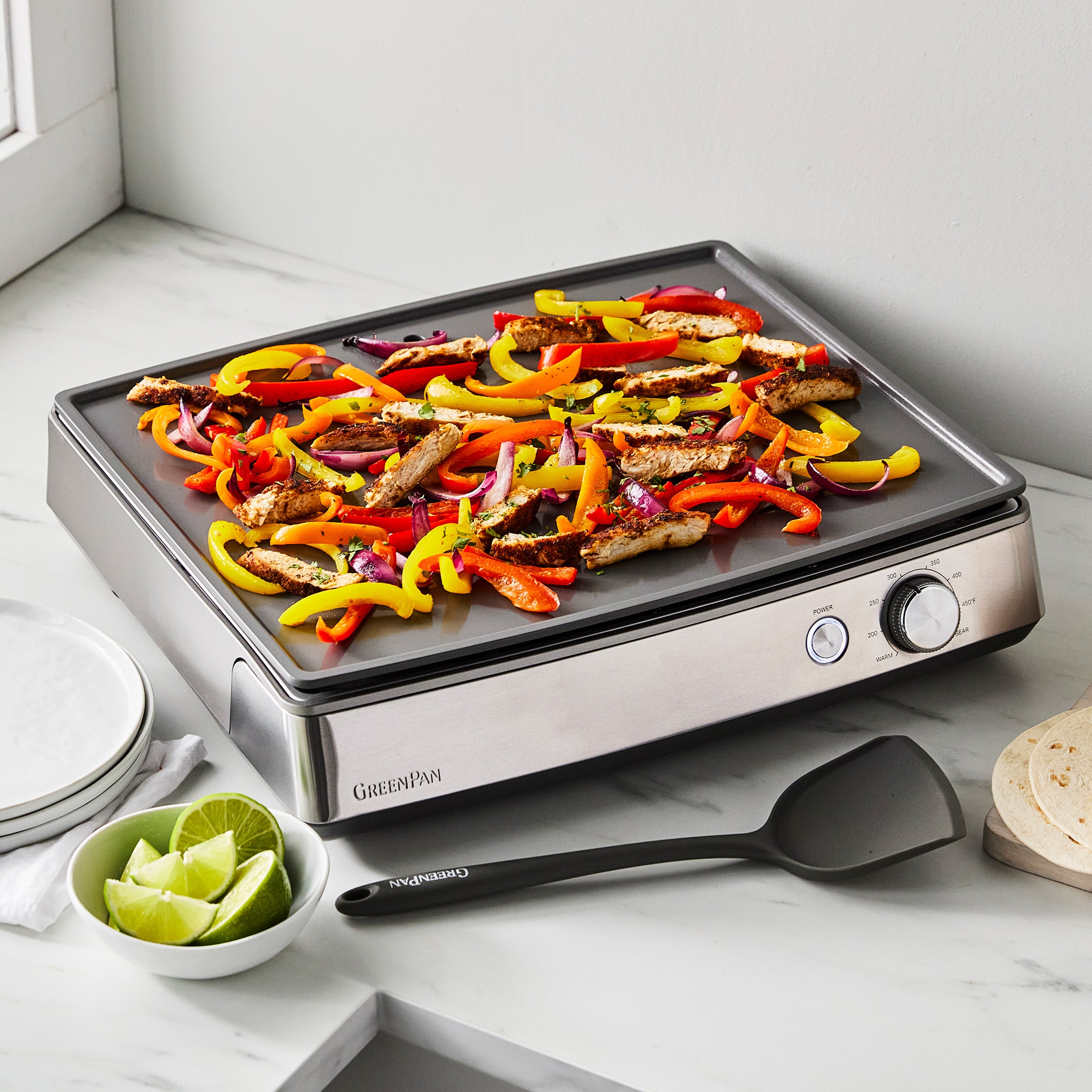 Stainless steel countertop griddle with chicken strips, peppers, lime wedges, spatula, and plates on a marble counter.