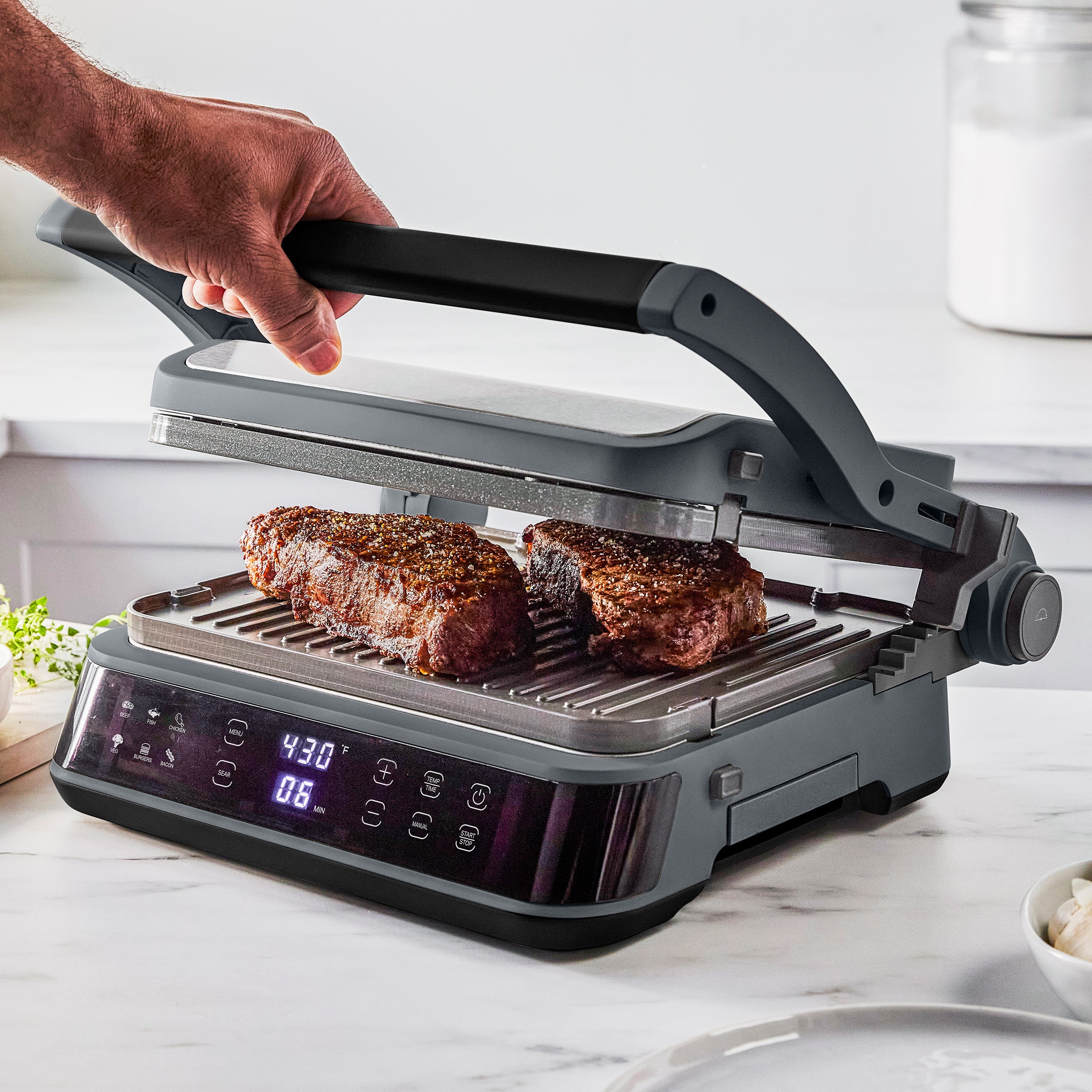 A hand closes the graphite-colored grill lid as two thick steaks cook on the kitchen counter.