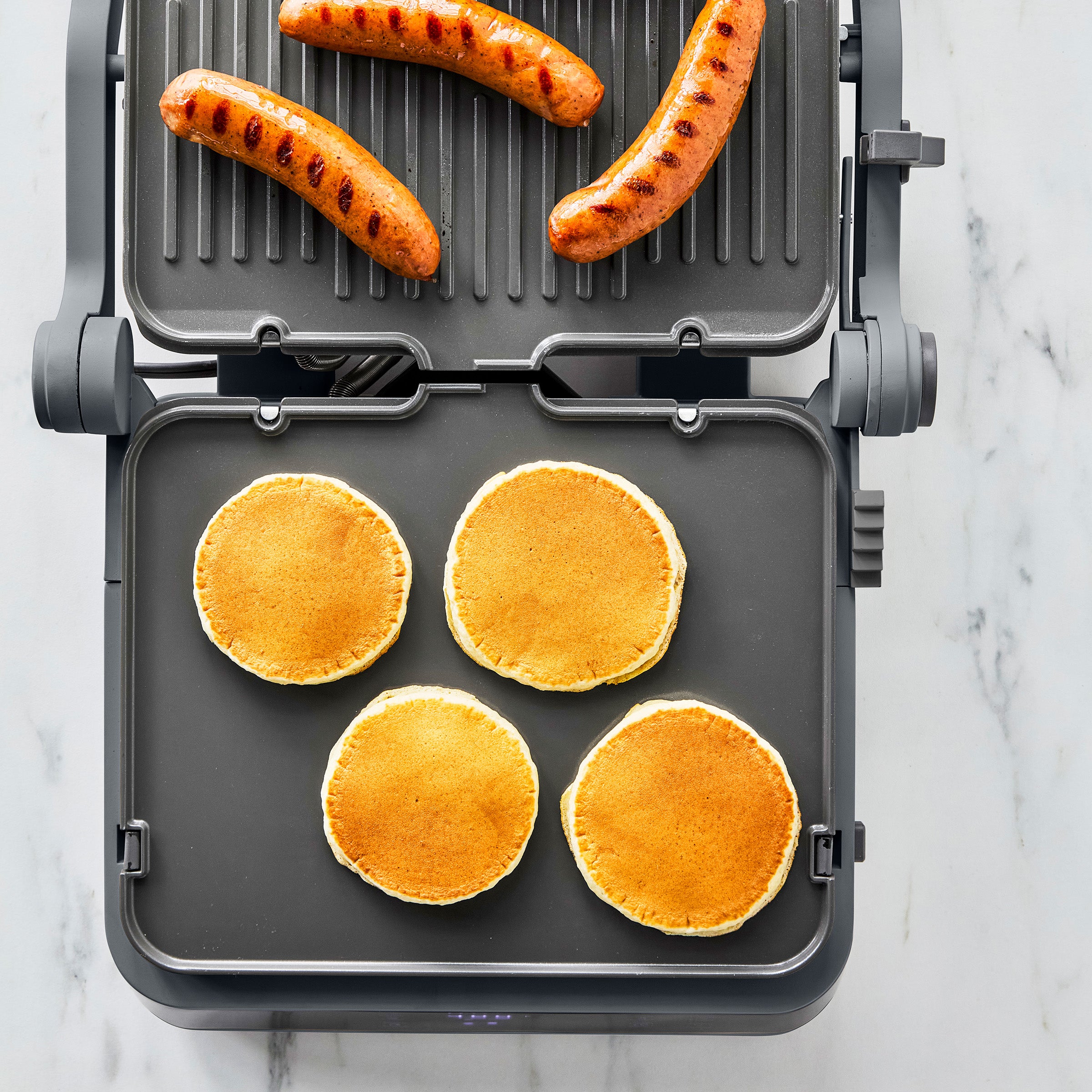 Four pancakes and three sausages cooking on a graphite-colored grill and griddle.