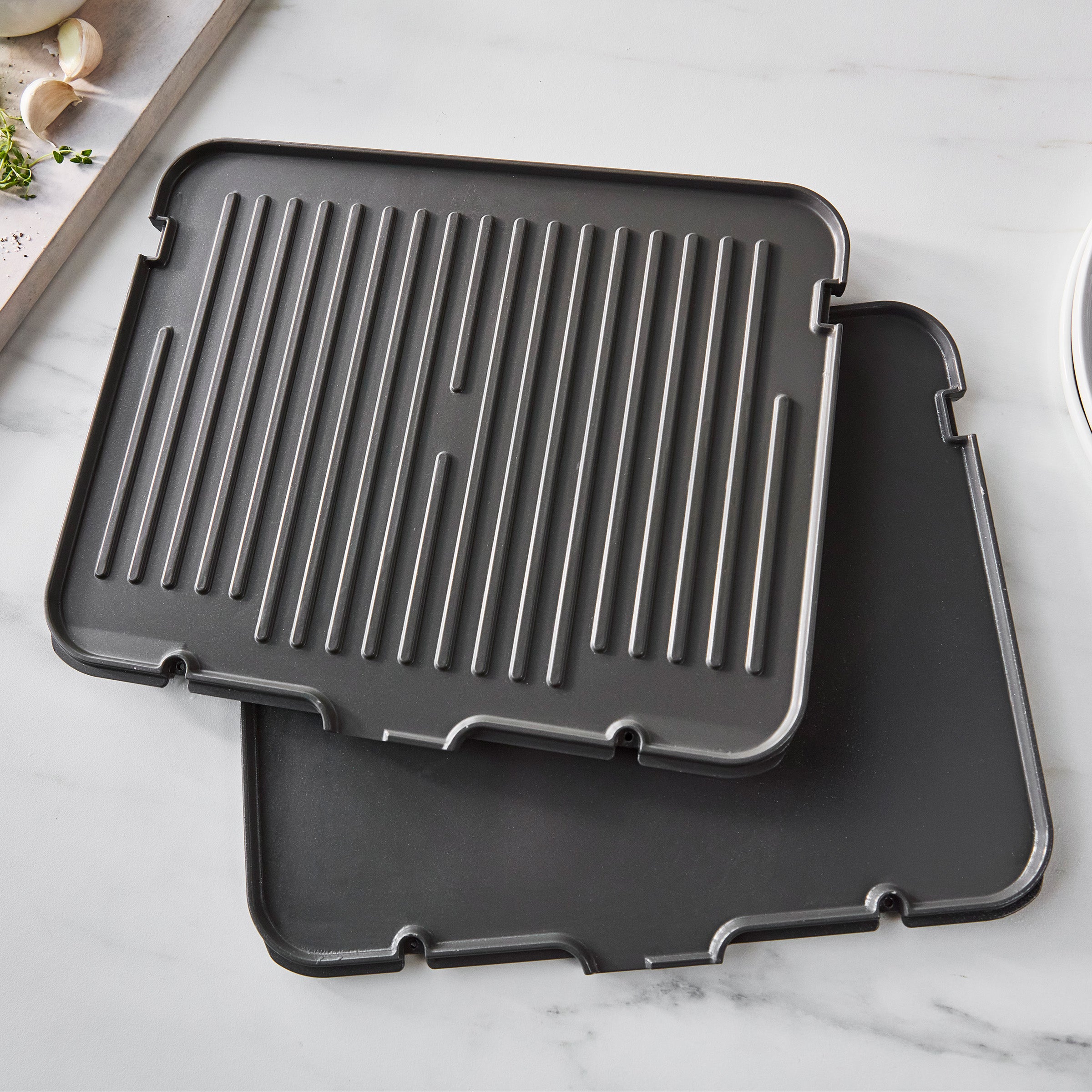 Two clay grill and griddle plates, one flat, one ridged, stacked on marble next to herbs and garlic.