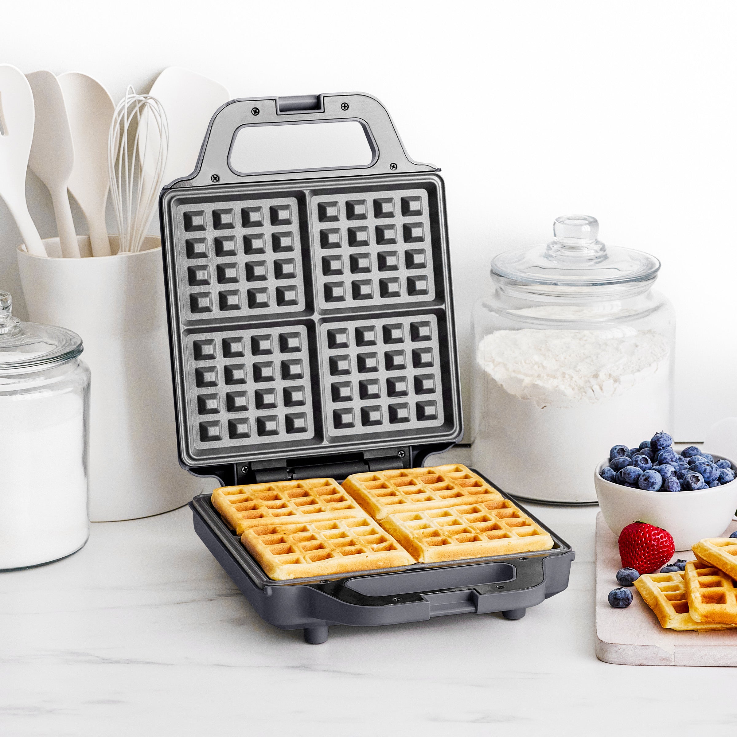 Waffle maker with waffles on a kitchen counter with utensils and ingredients.