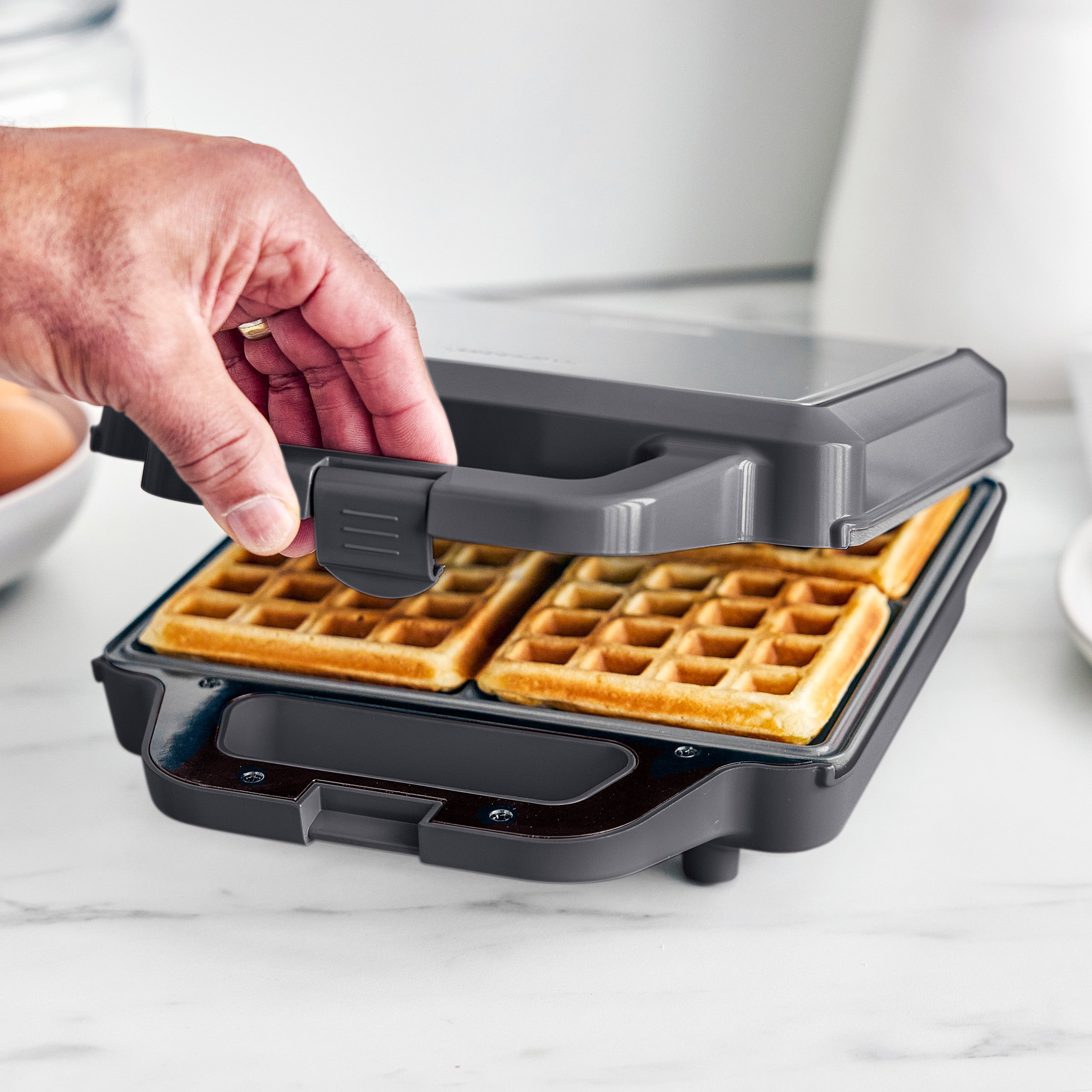 Person opening a waffle maker with golden brown waffles inside on a white surface.