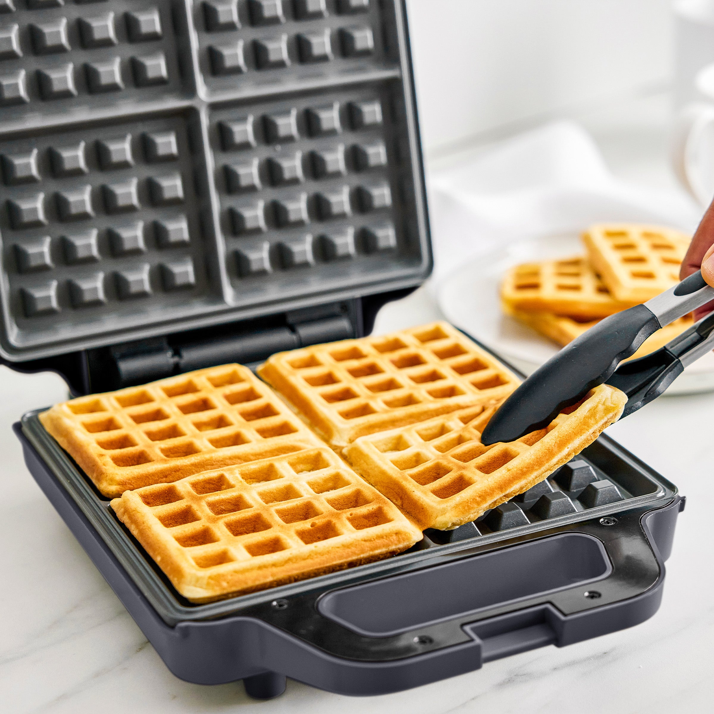 Waffle maker with freshly made waffles and a pair of tongs on a white surface.