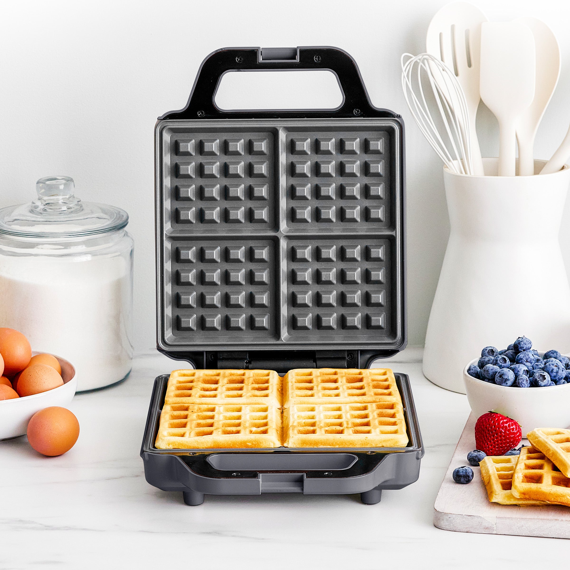Waffle maker with waffles on a kitchen counter with ingredients and utensils.