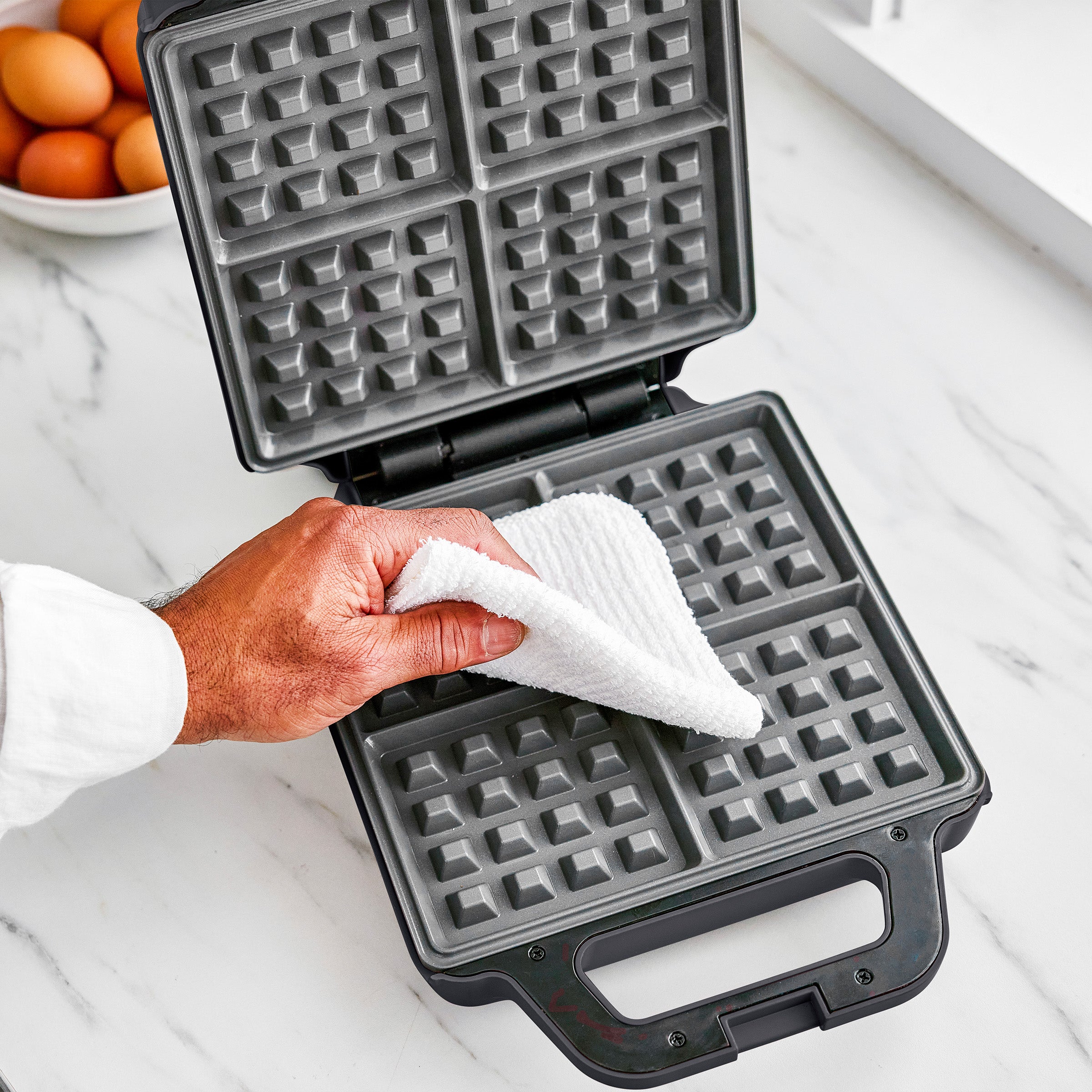 Person cleaning a waffle maker with a white cloth on a marble countertop.